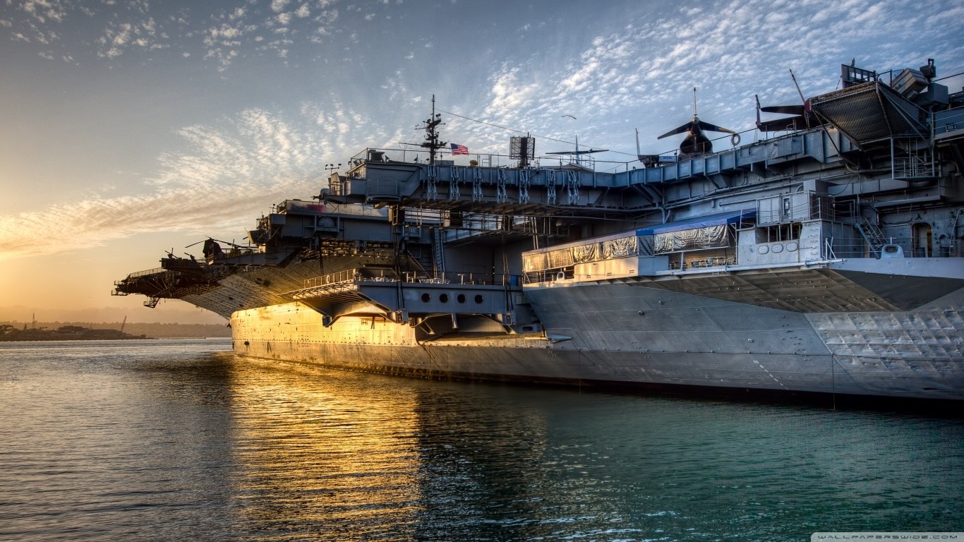 USS Midway (CV-41) aircraft carrier docked at sunset, reflecting golden light on the water with a partly cloudy sky above.