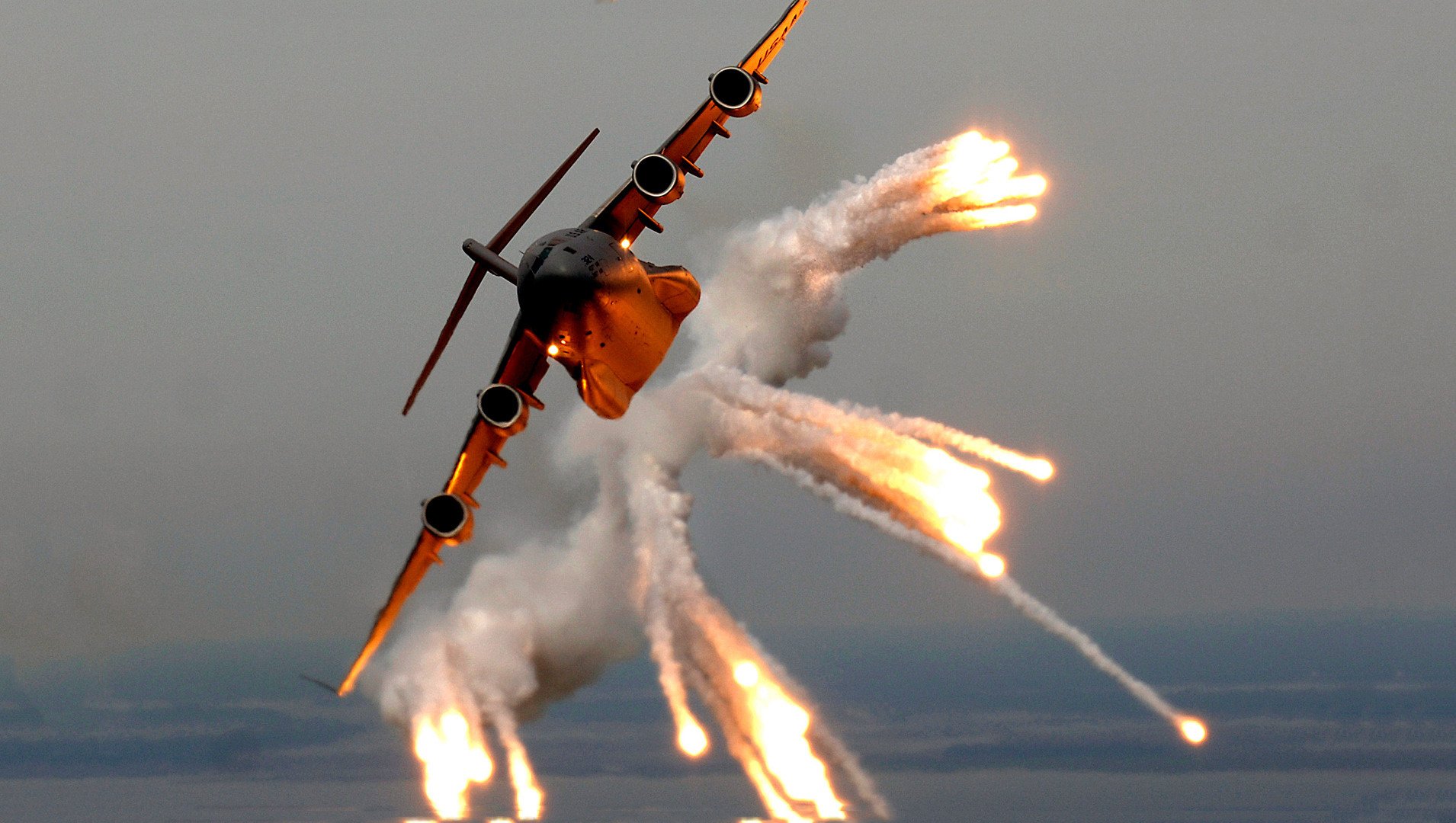 Military Boeing C-17 Globemaster III banking sharply while releasing bright countermeasure flares over the sea.