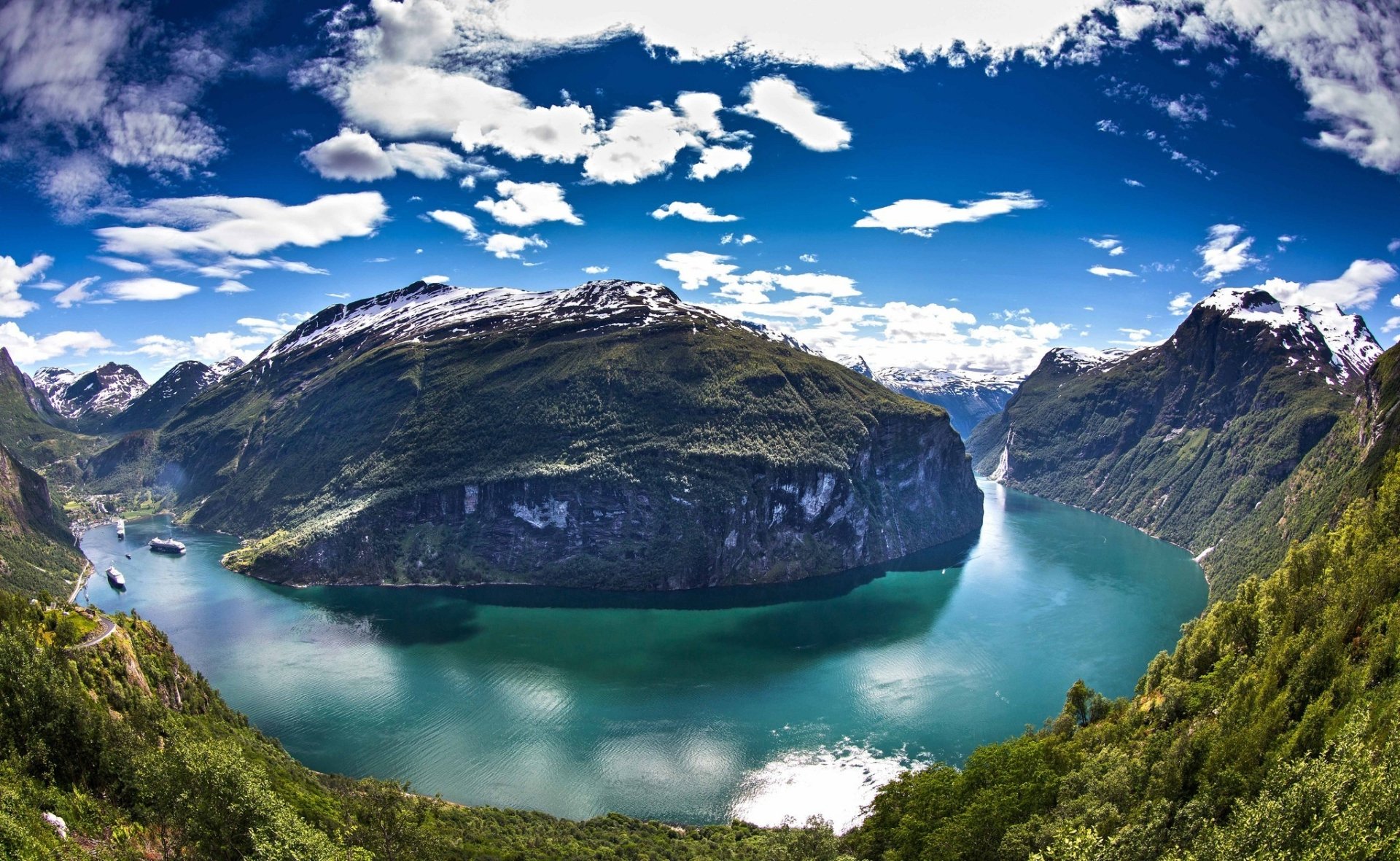 Norway fjord landscape: turquoise water winding through steep green mountains and snow‑capped peaks, a waterfall and vivid blue sky — a dramatic nature scene.
