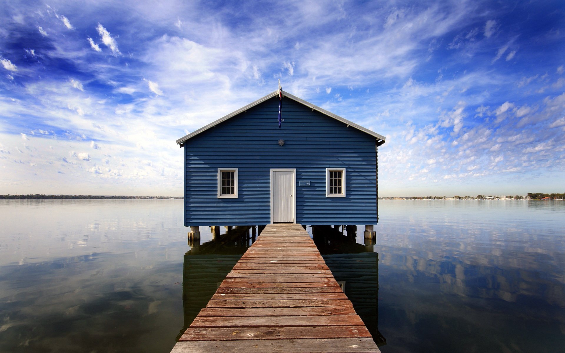 A serene blue boathouse stands on stilts over calm water, with a wooden pier leading out to it, under a sky adorned with soft clouds.