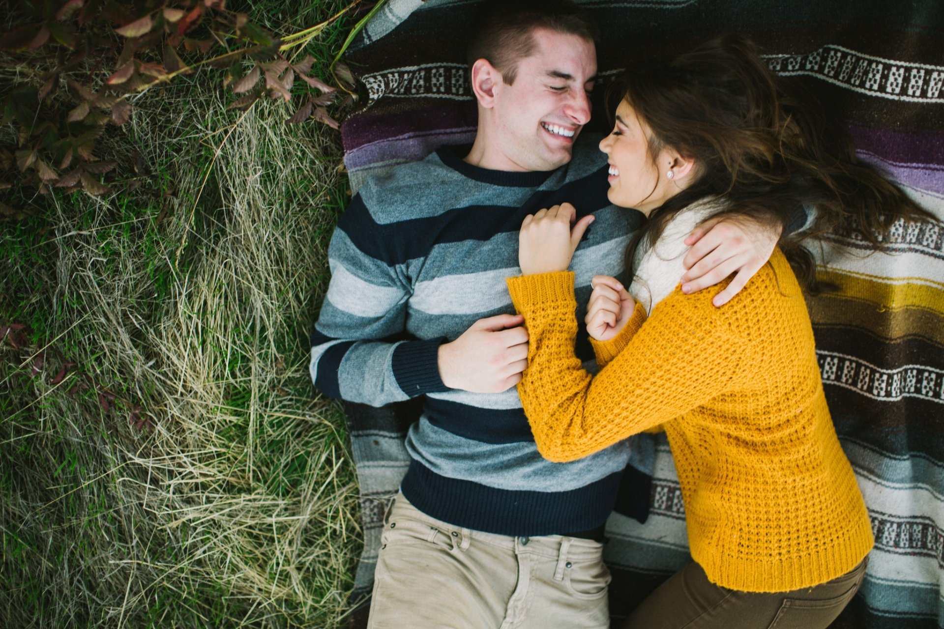 A couple lies on a colorful blanket, laughing and gazing into each other's eyes. Their cozy embrace captures a moment of love and connection in a natural setting.