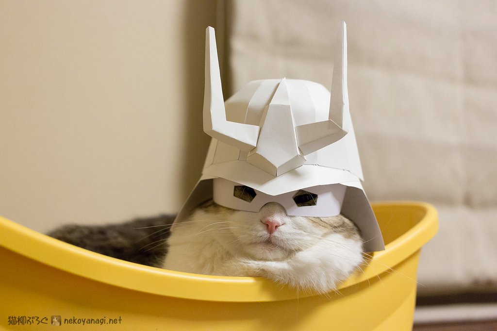 A playful cat wearing a paper hat styled like a samurai helmet, resting comfortably in a yellow container.