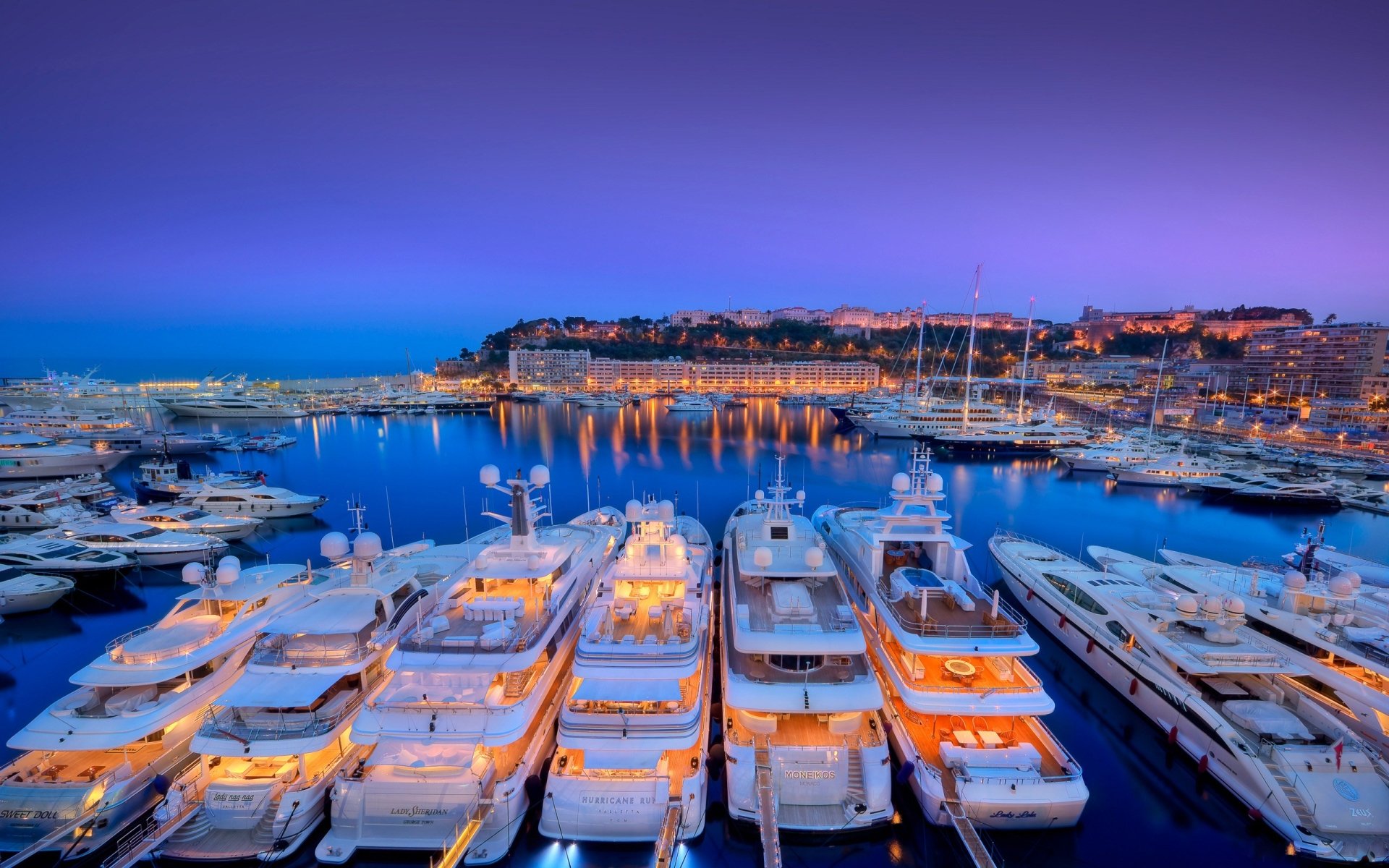 HDR image of Monaco’s harbor at sunset, showcasing yachts docked in the man-made marina with the city and ocean in the background under a vibrant sky.