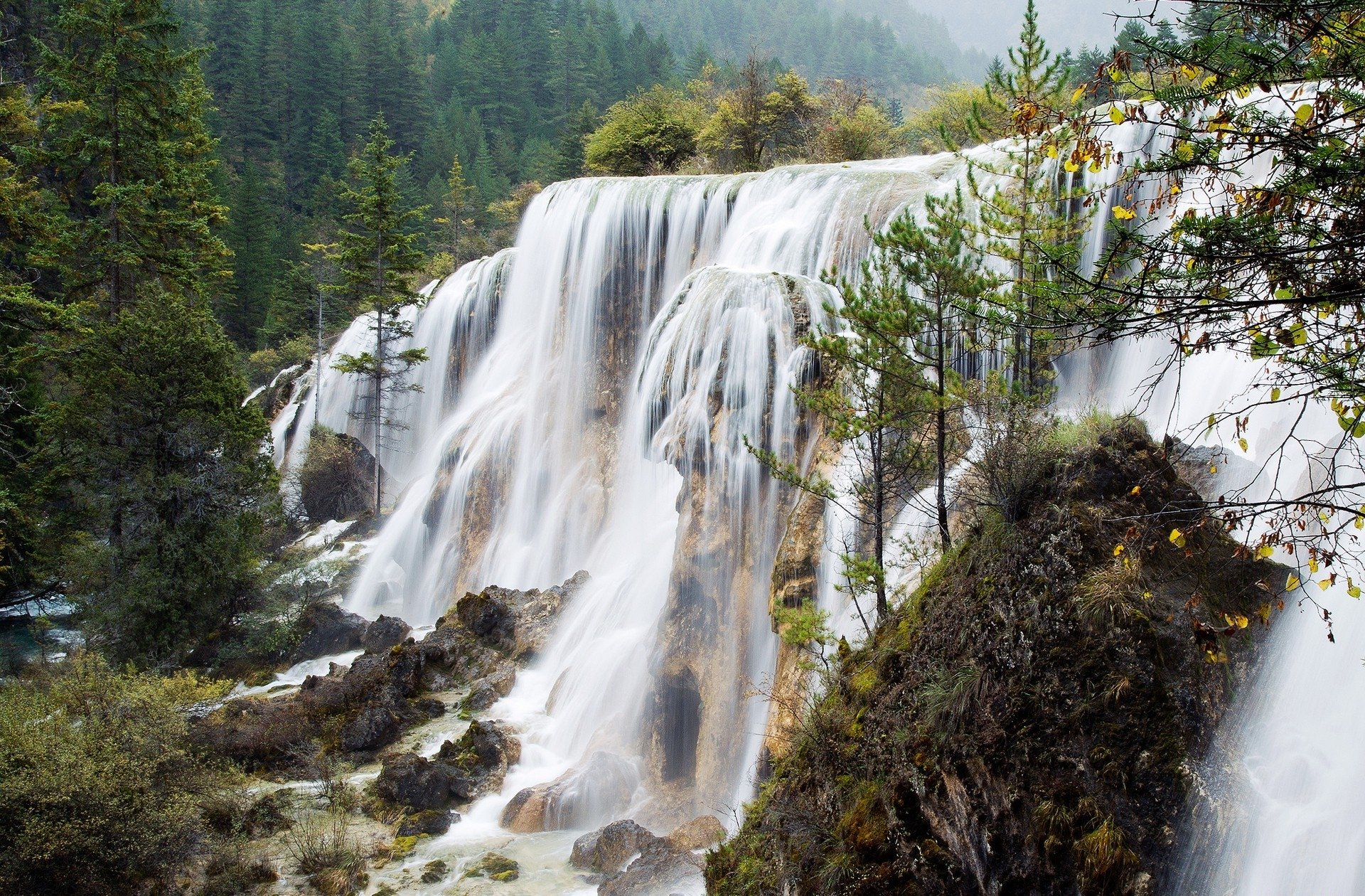 Tiered waterfall cascading over rocky terraces through evergreen forest at Jiuzhaigou Park, China — a Tibetan-region mountain nature scene.