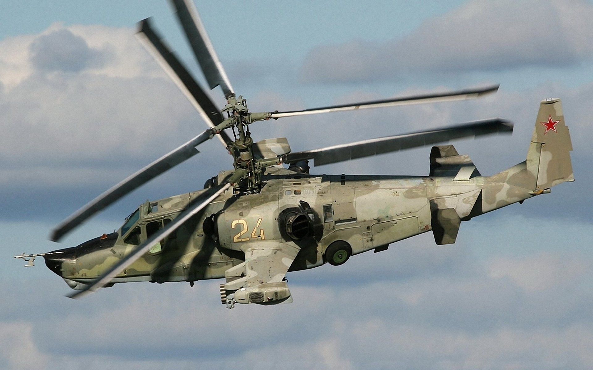 A military Kamov Ka-50 helicopter in flight with visible twin coaxial rotors and camouflage paint against a cloudy sky backdrop.