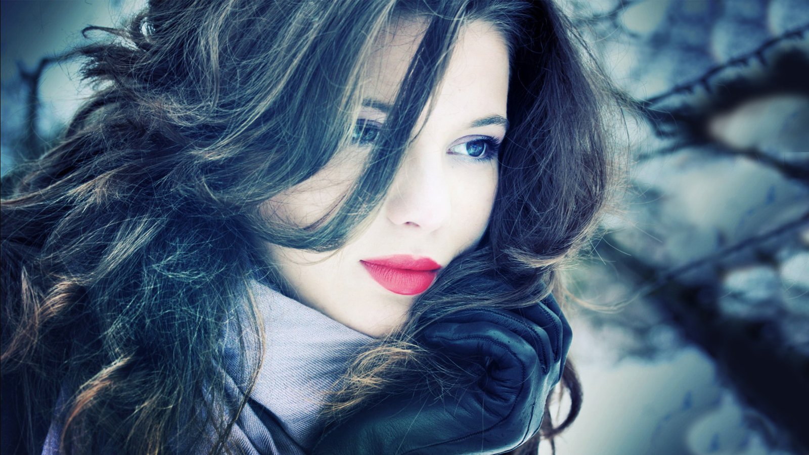 A woman with striking features gazes thoughtfully, her long, dark hair framing her face. She wears a cozy scarf and gloves, set against a softly blurred winter backdrop.