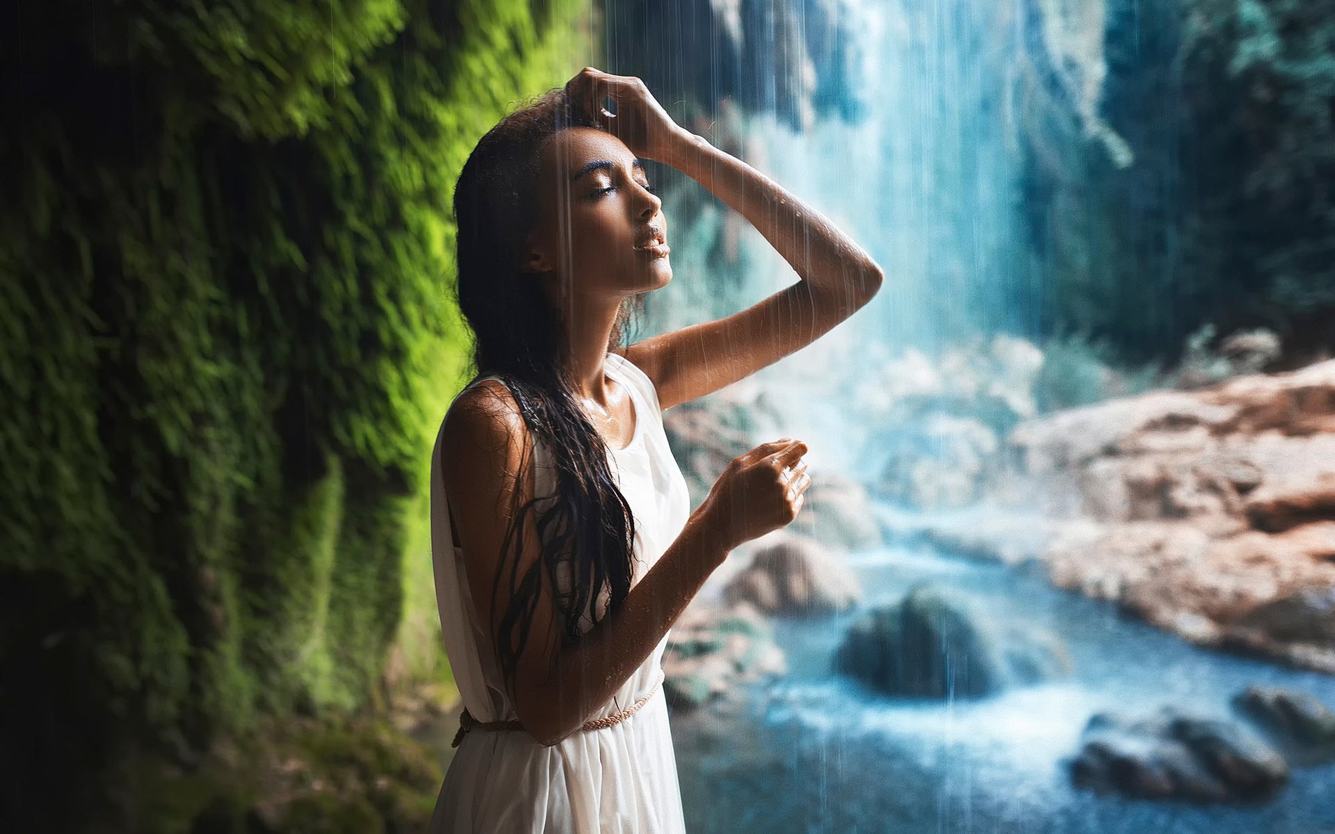 A woman in a white dress stands near a waterfall, her eyes closed and hand resting on her forehead, reflecting a serene and contemplative mood.