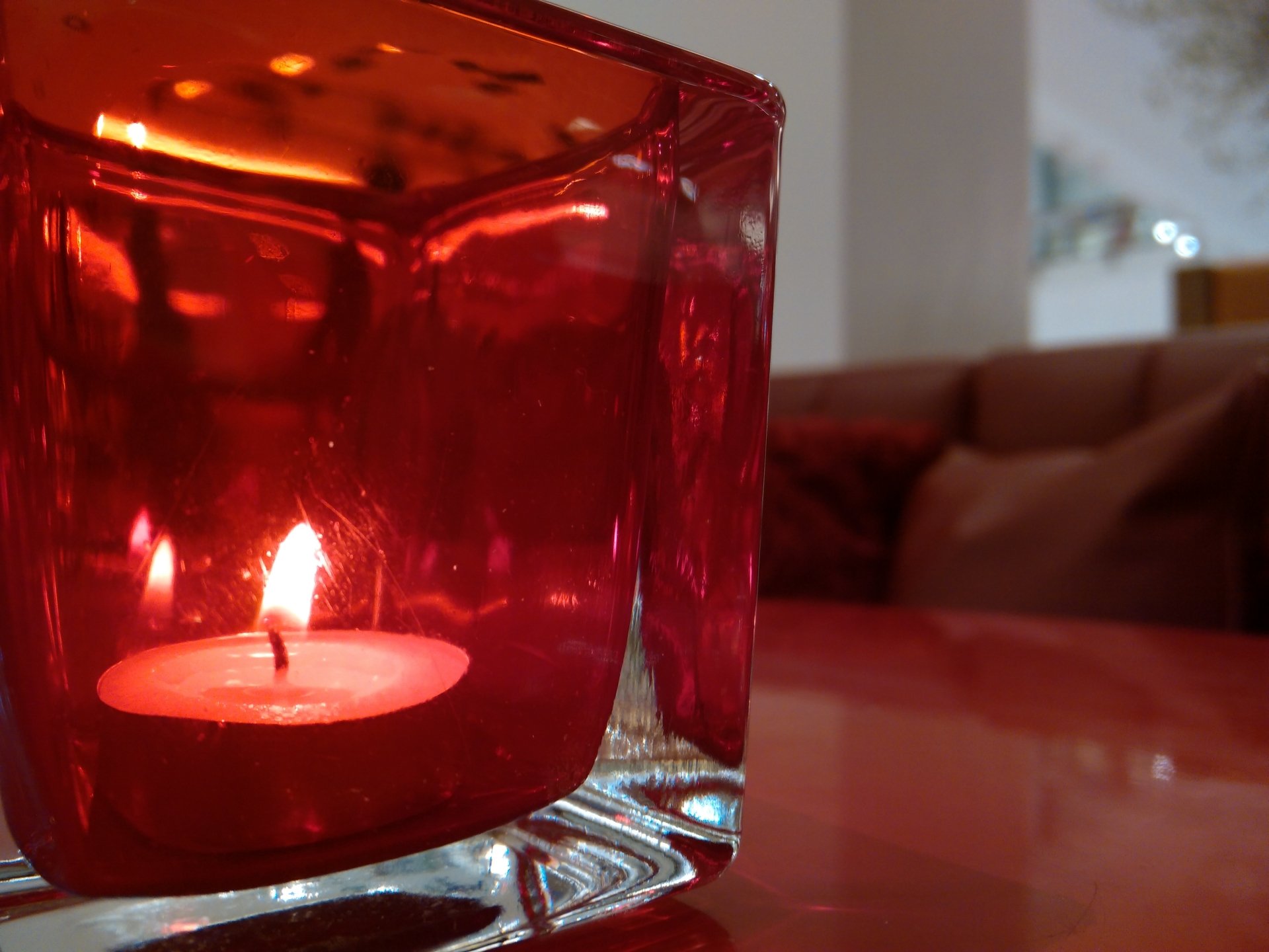Photography of a lit tealight candle in a red glass holder on a glossy table, warm living room sofa softly blurred in the background.