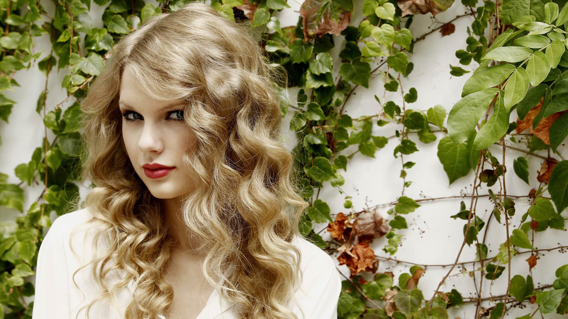A close-up portrait of Taylor Swift with wavy blonde hair, set against a backdrop of green ivy, exuding a confident and vibrant musical presence.