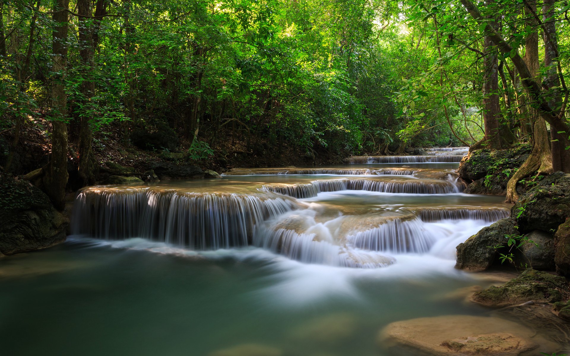 A peaceful river flows gently over layered rocks surrounded by lush green trees in a serene natural setting.