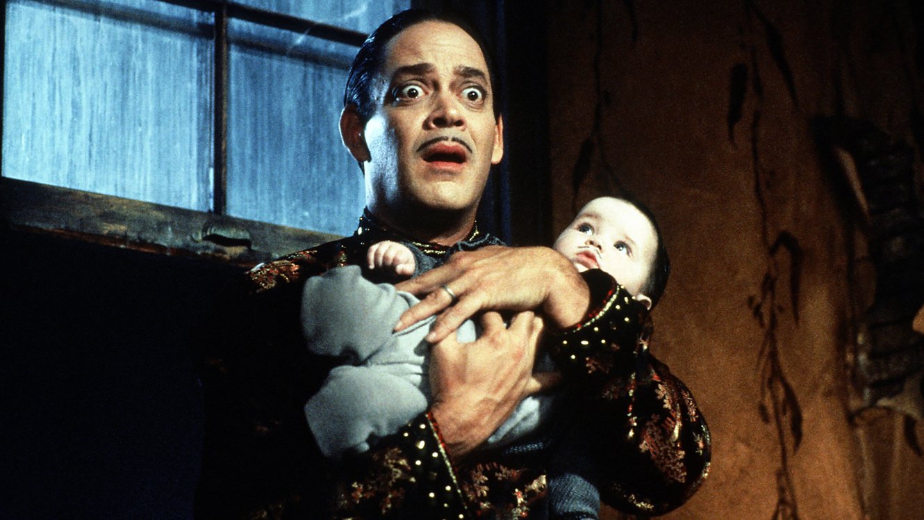 Man holding a baby indoors with a worried expression in a scene from the movie Addams Family Values.