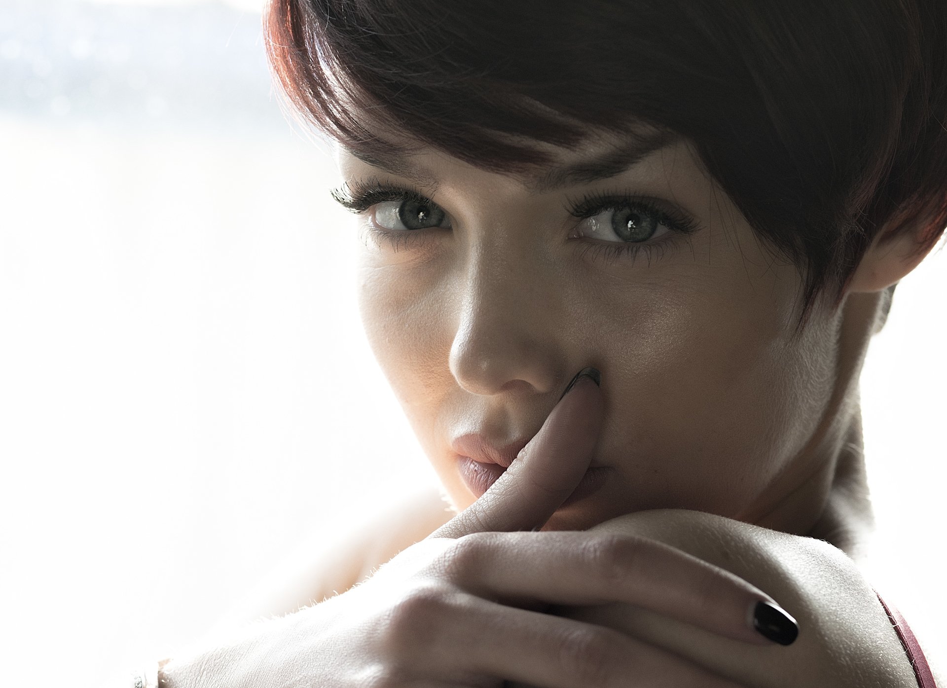 Close-up of a brunette woman in a moody, contemplative pose, fingers near her lips and a soft, intense gaze.
