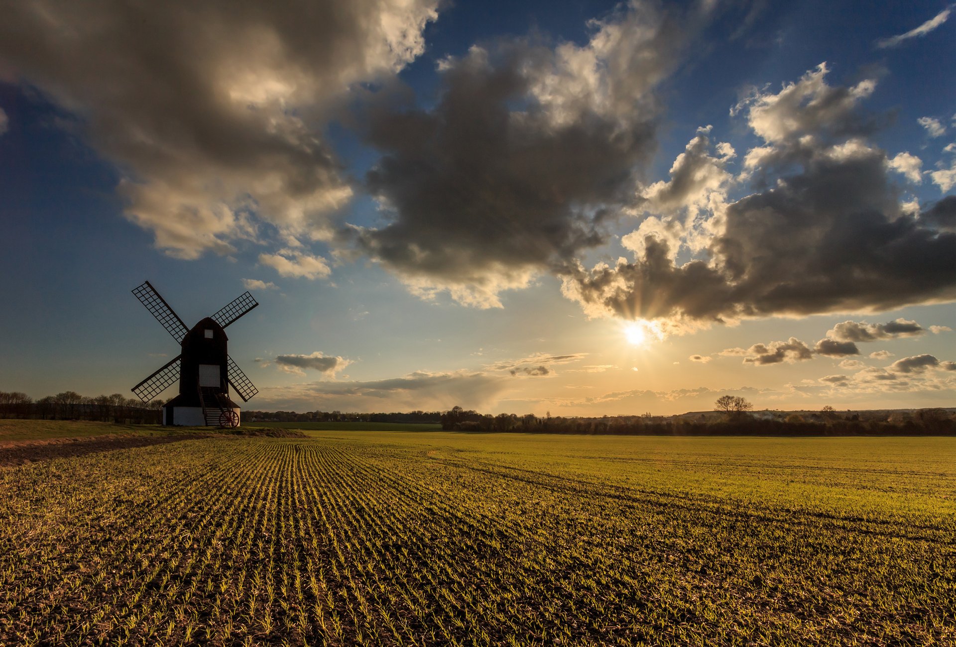 Man-made windmill stands at the edge of a plowed field while the low sun breaks through dramatic clouds.