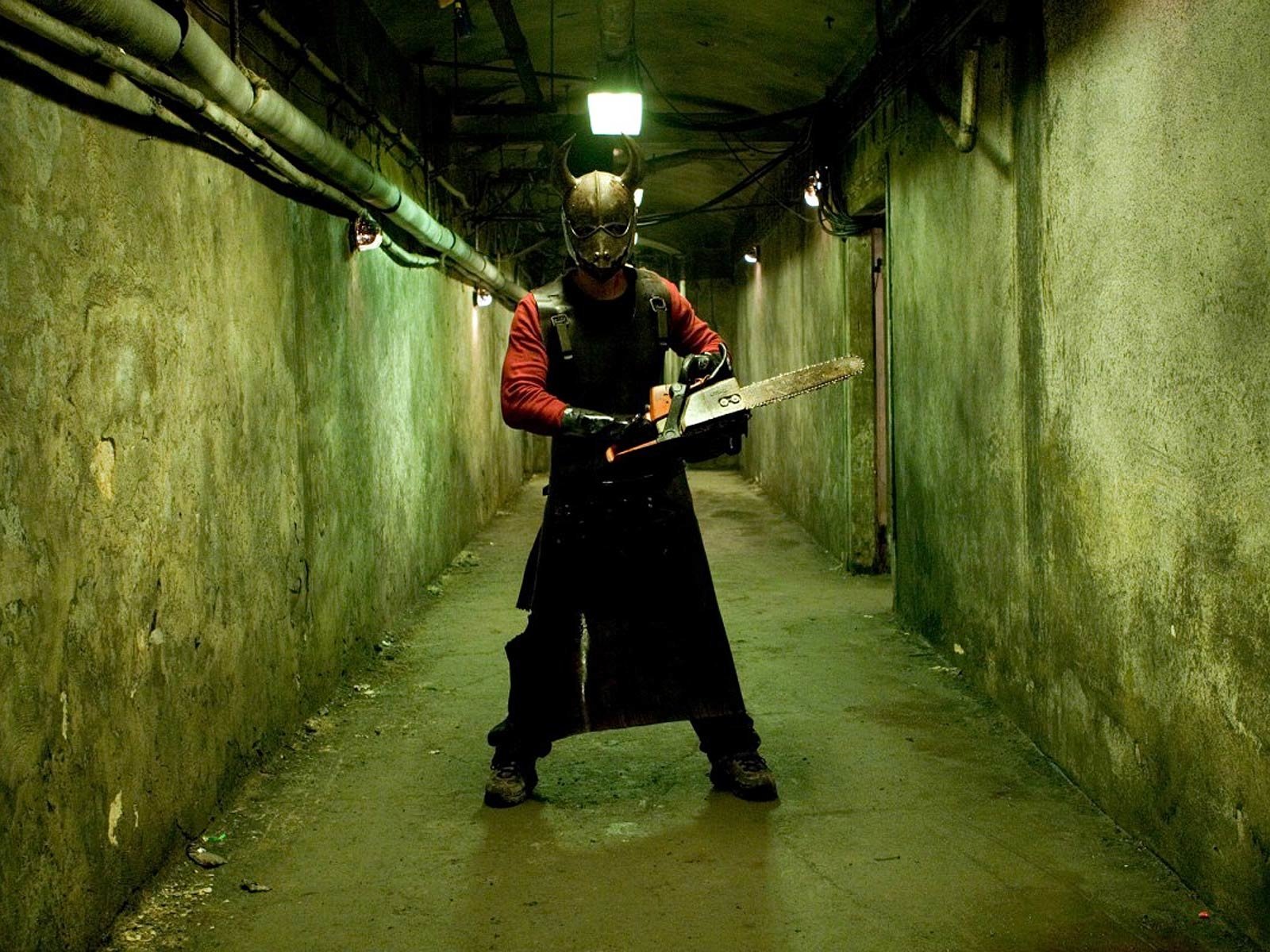Movie scene in a grim hostel corridor: a masked, apron-clad attacker stands under a single light wielding a chainsaw, grimy concrete walls and exposed pipes.