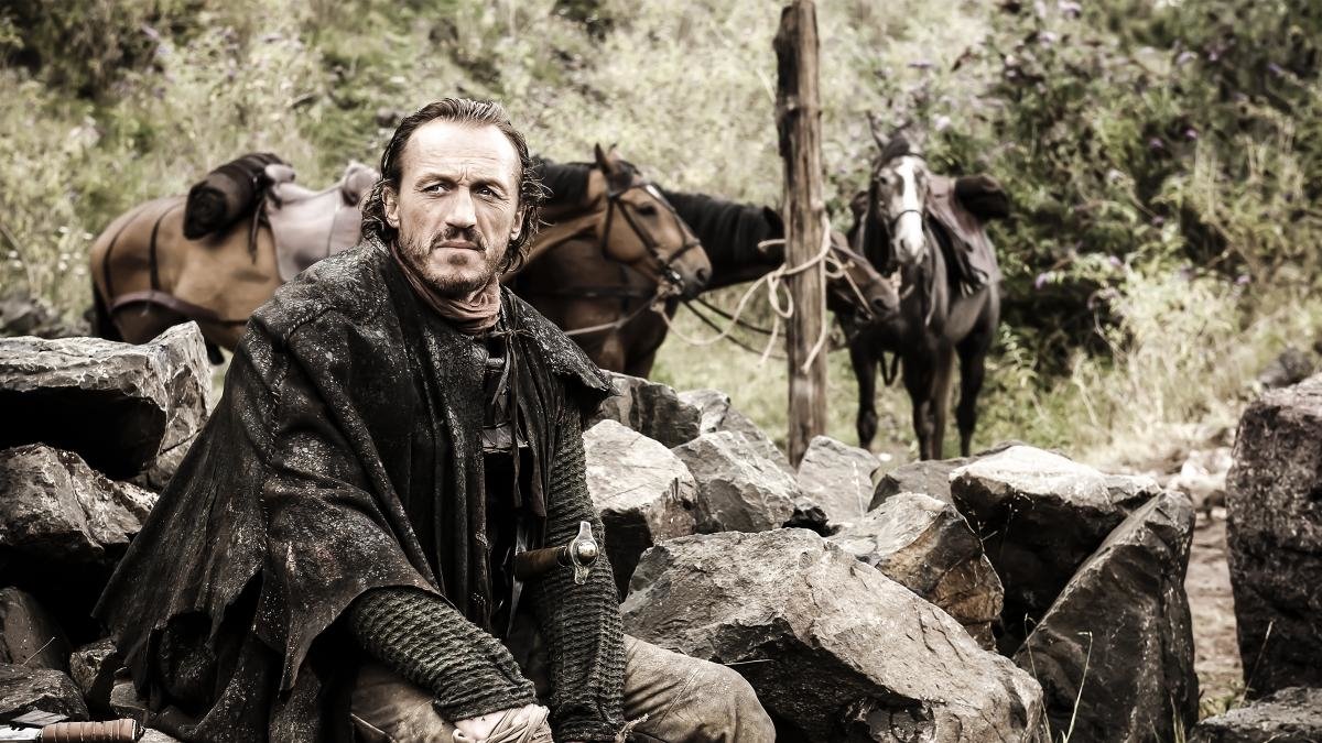 A brooding character from the TV show Game of Thrones sits among rocks, set against a backdrop of grazing horses and rugged terrain.