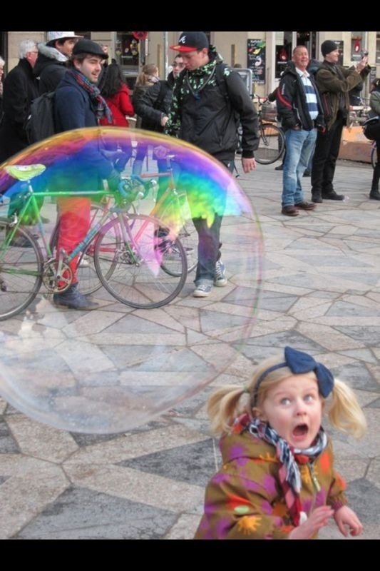 A child with a surprised expression watches a giant colorful bubble float near bicycles, capturing a funny and unexpected moment in a busy outdoor setting.