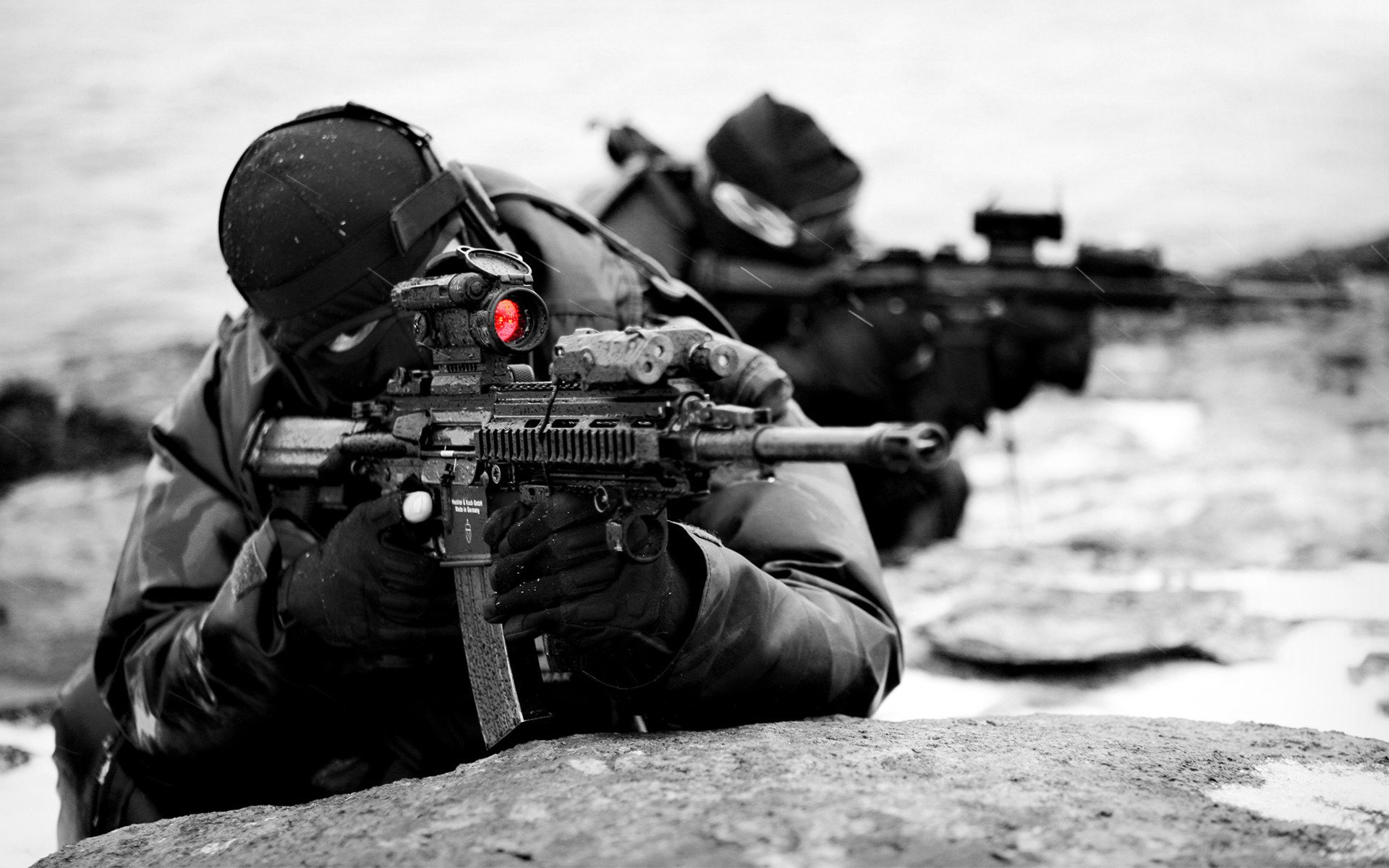 Two military soldiers in tactical gear aim rifles while lying prone behind rocks in a black-and-white setting.