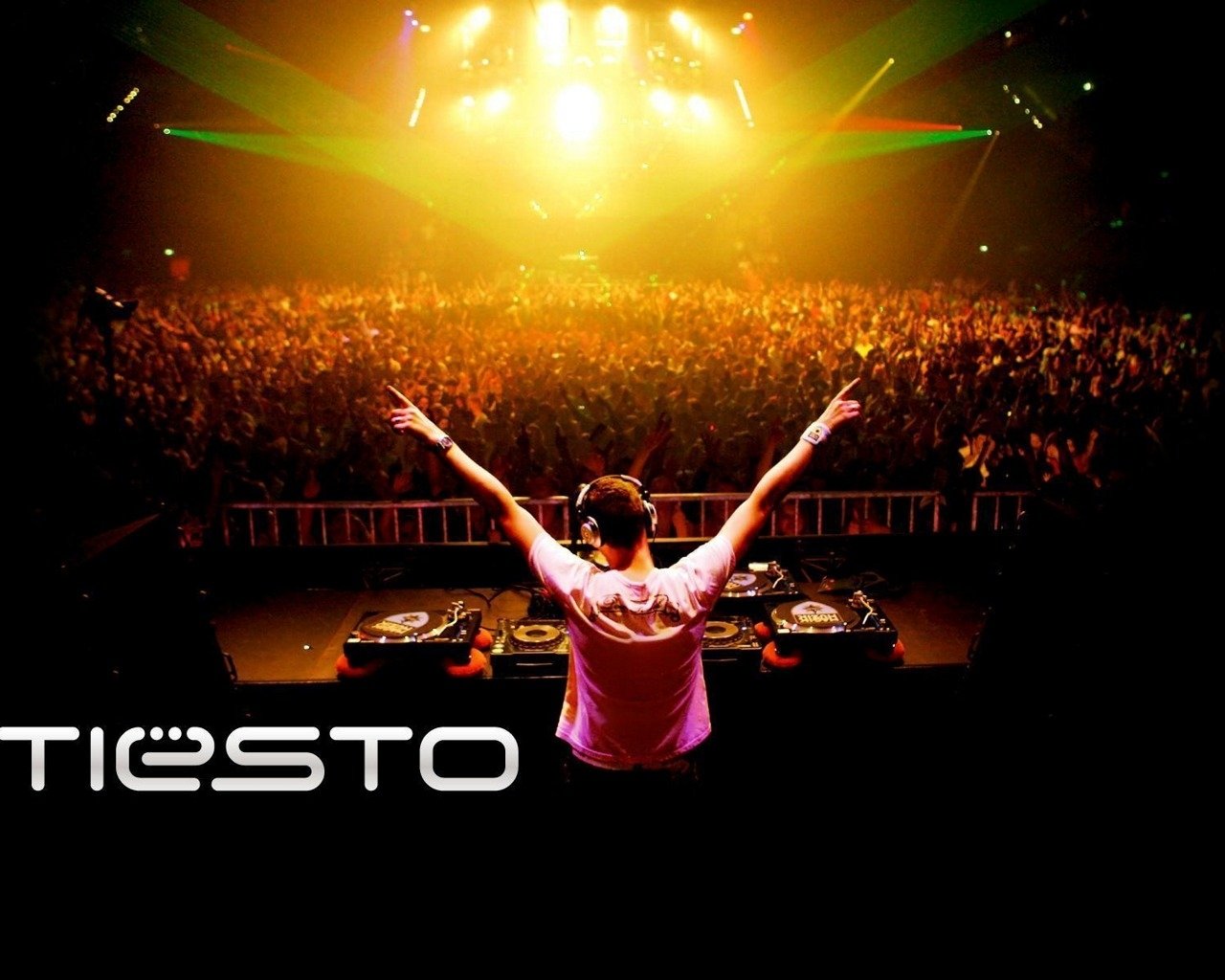 A DJ performs energetically on stage, arms raised, in front of a vibrant, cheering crowd, with colorful lights illuminating the venue, capturing the excitement of a live music event.