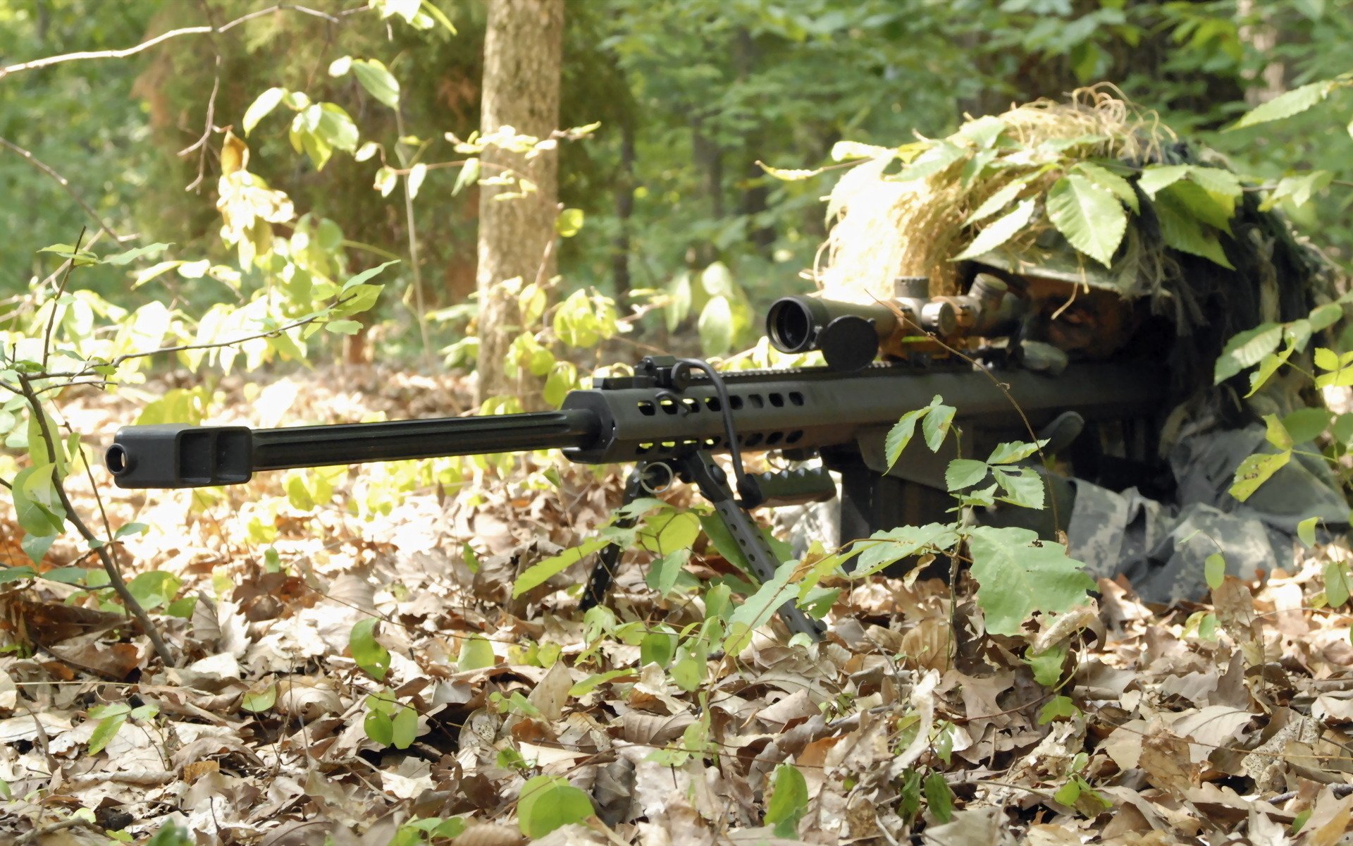 A military sniper lies camouflaged among leaves, poised with a rifle and focused on the surroundings, surrounded by a wooded environment.