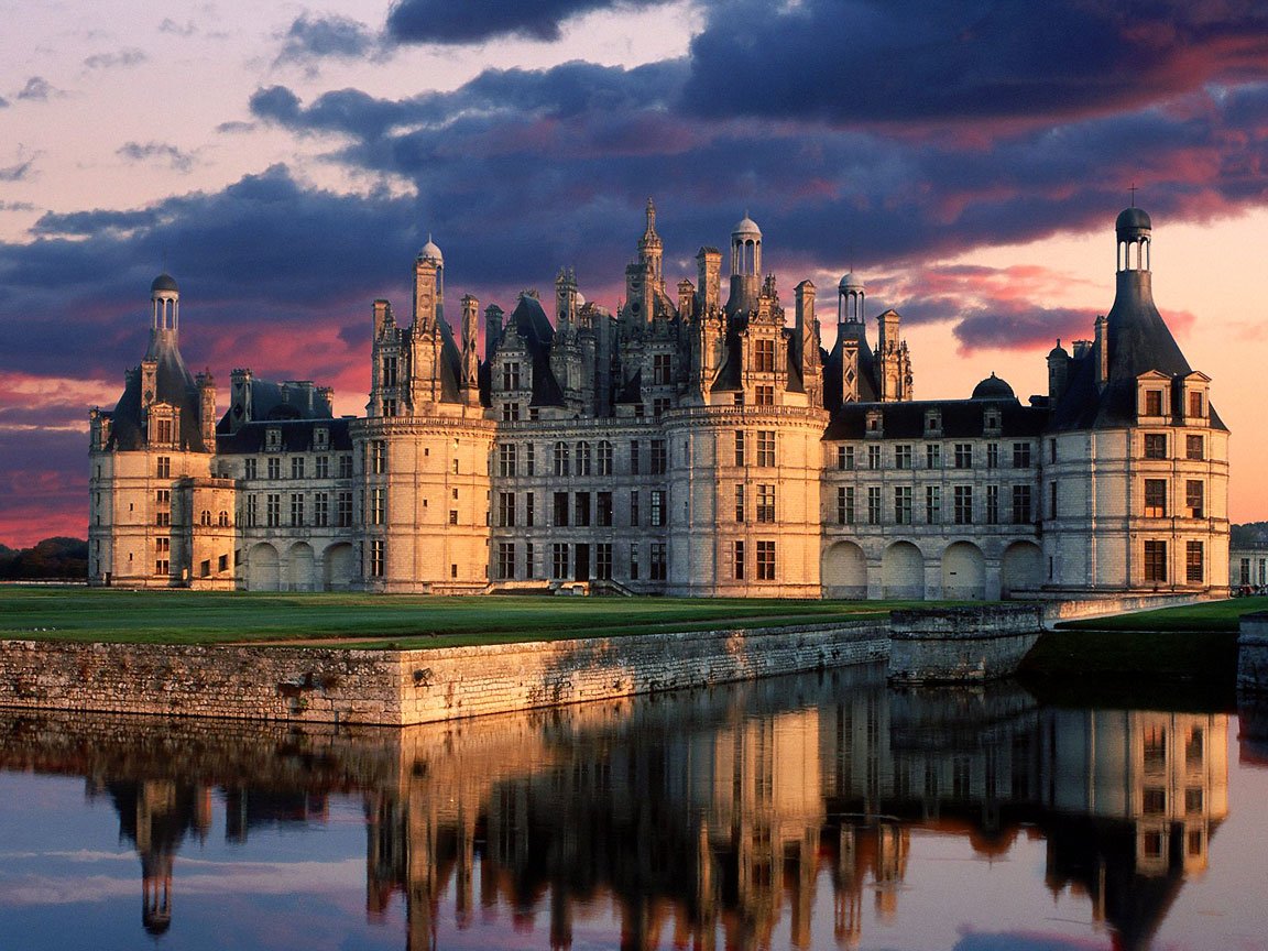 Château de Chambord, a grand man-made Renaissance castle, stands reflected in the calm water at sunset under a dramatic sky.