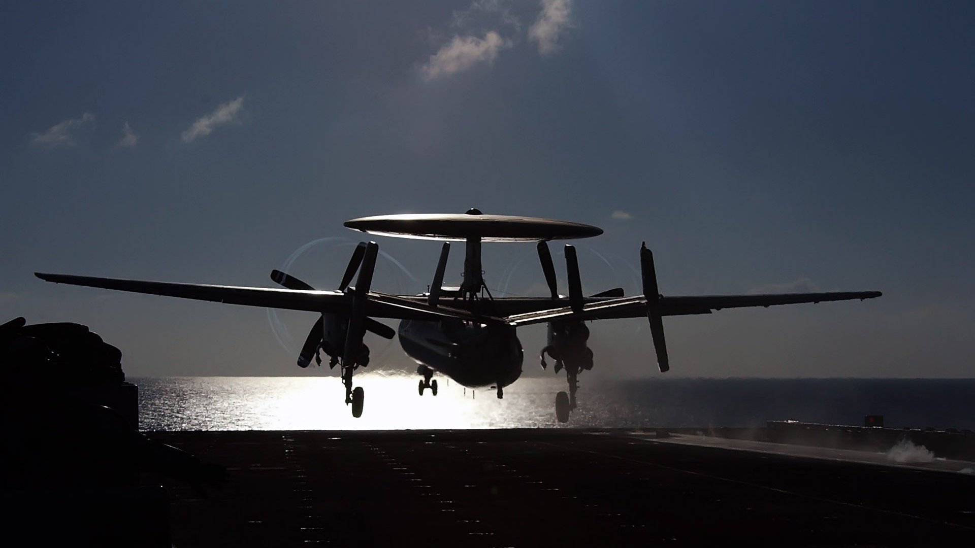 Silhouetted military Northrop Grumman E-2 Hawkeye with radar dome landing on an aircraft carrier deck at dusk, ocean and sky glowing behind it.