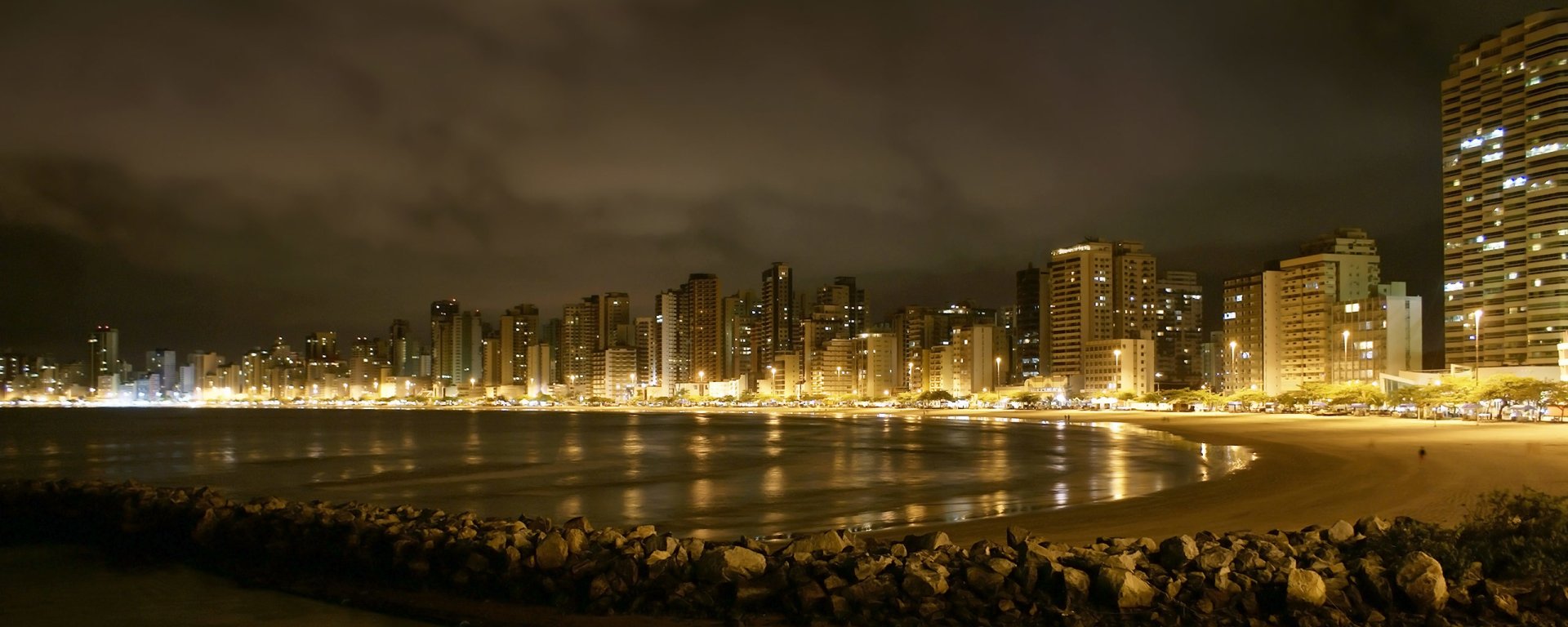 Nighttime view of a man-made cityscape along a curved shoreline, with illuminated buildings reflecting off the calm water.