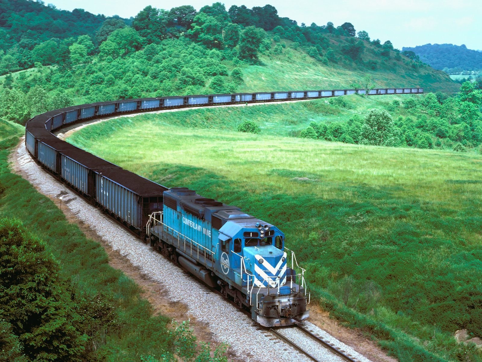 A blue and silver train navigates a winding track through lush green hills, pulling a long line of freight cars along a scenic landscape.