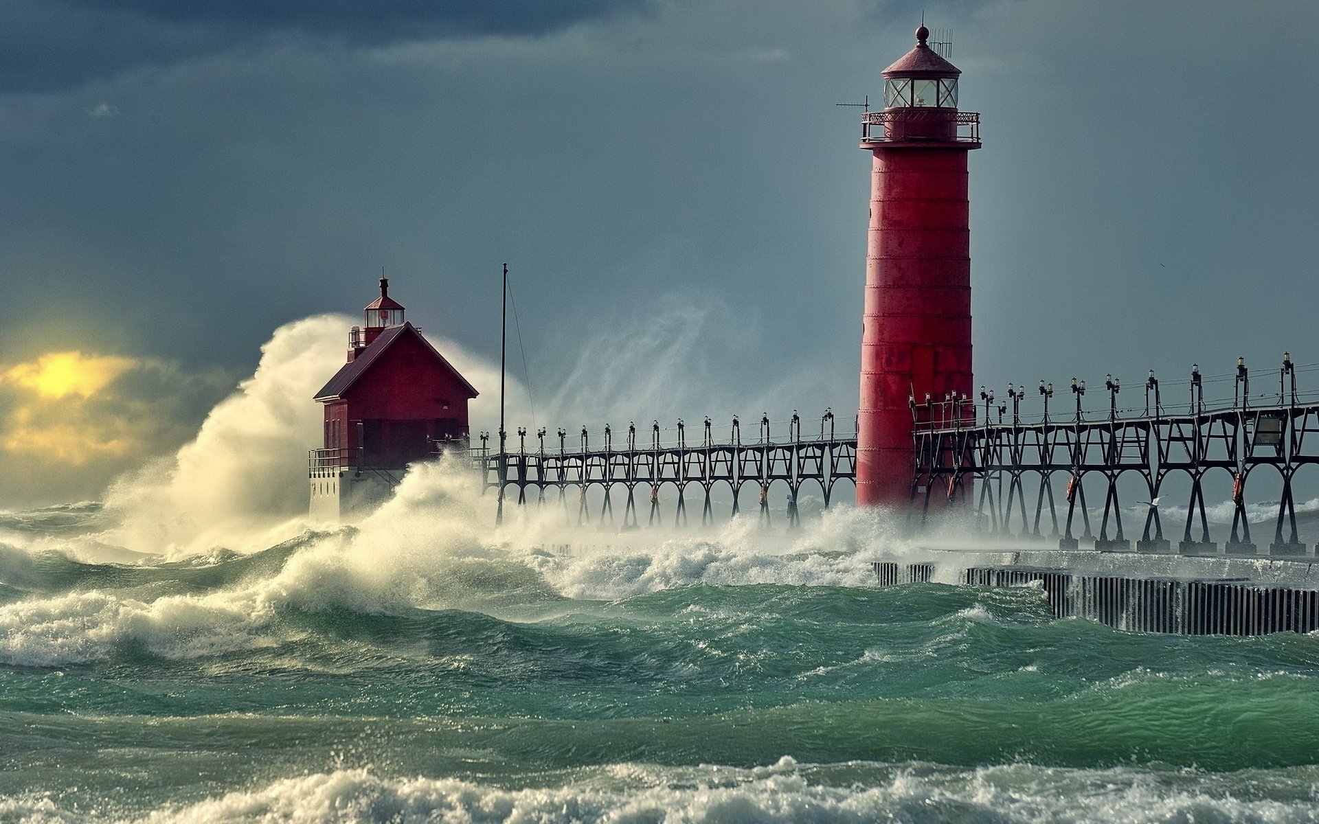 Man-made red lighthouse and keeper's house on a pier with towering waves crashing and spray under a stormy sky.