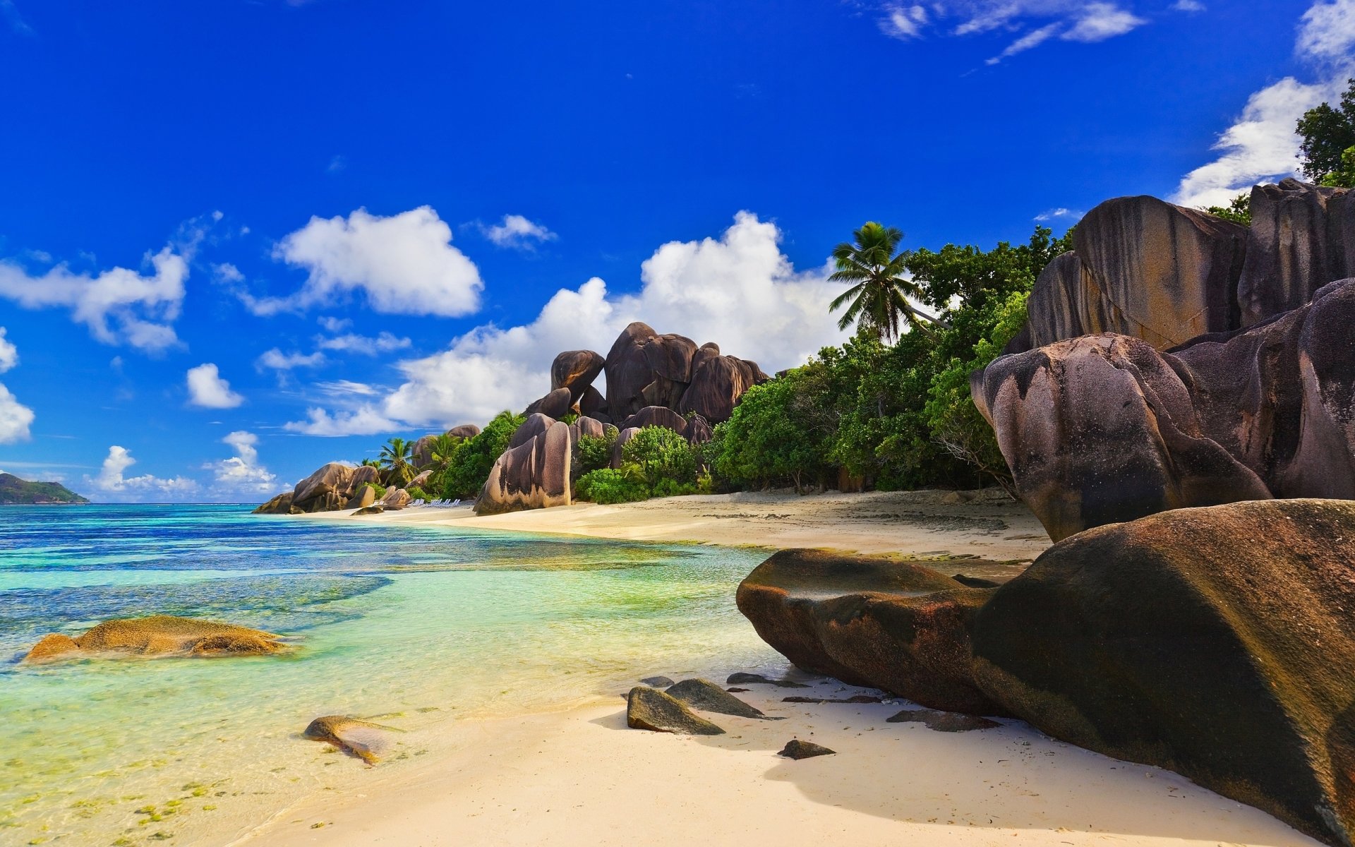 Nature beach scene: white sand shoreline, turquoise shallow sea, rounded granite boulders and palm trees beneath a vivid blue sky with scattered clouds.