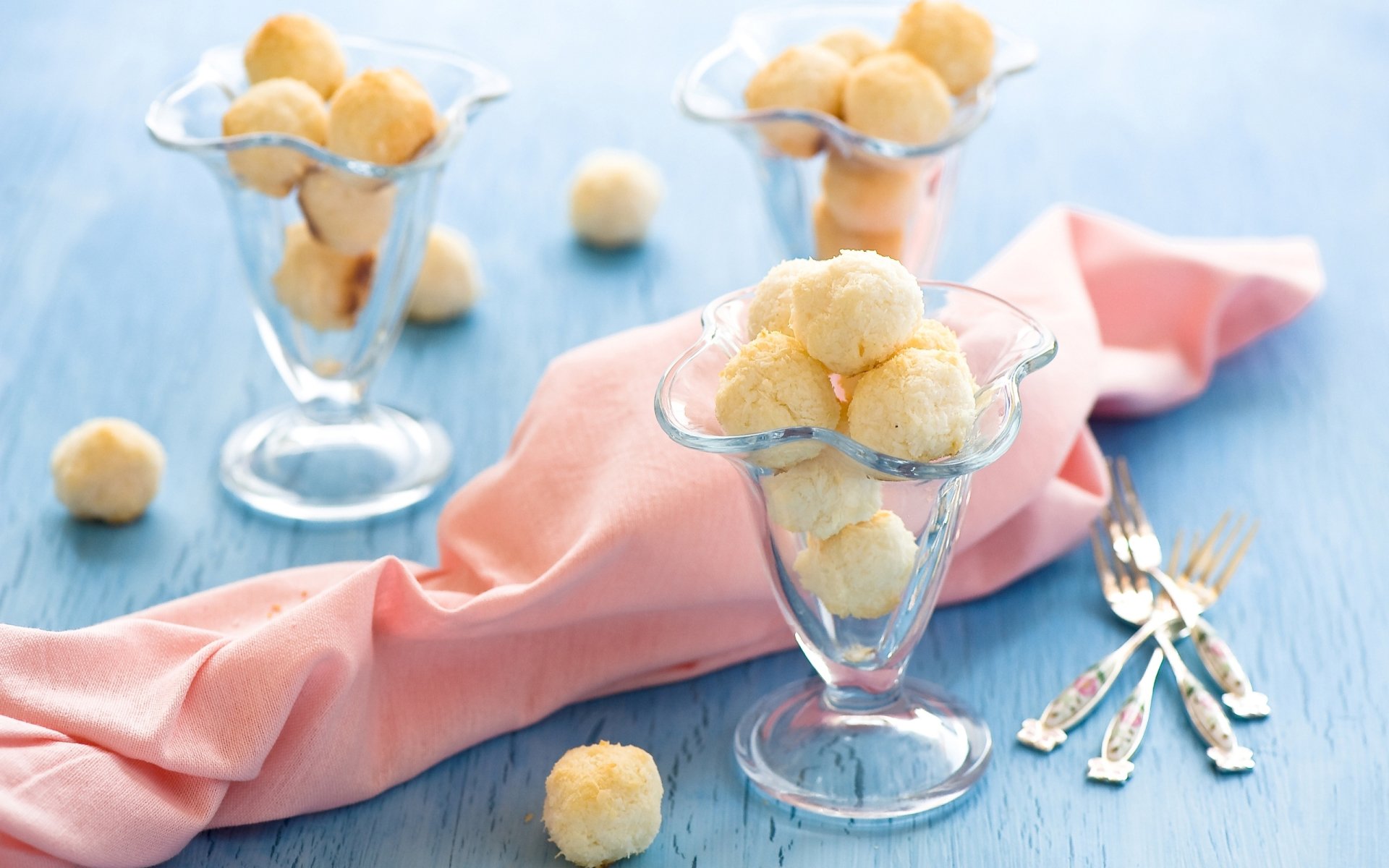Two glass dessert cups filled with small round cookies sit on a blue wooden surface with a pink cloth and silver dessert forks nearby.