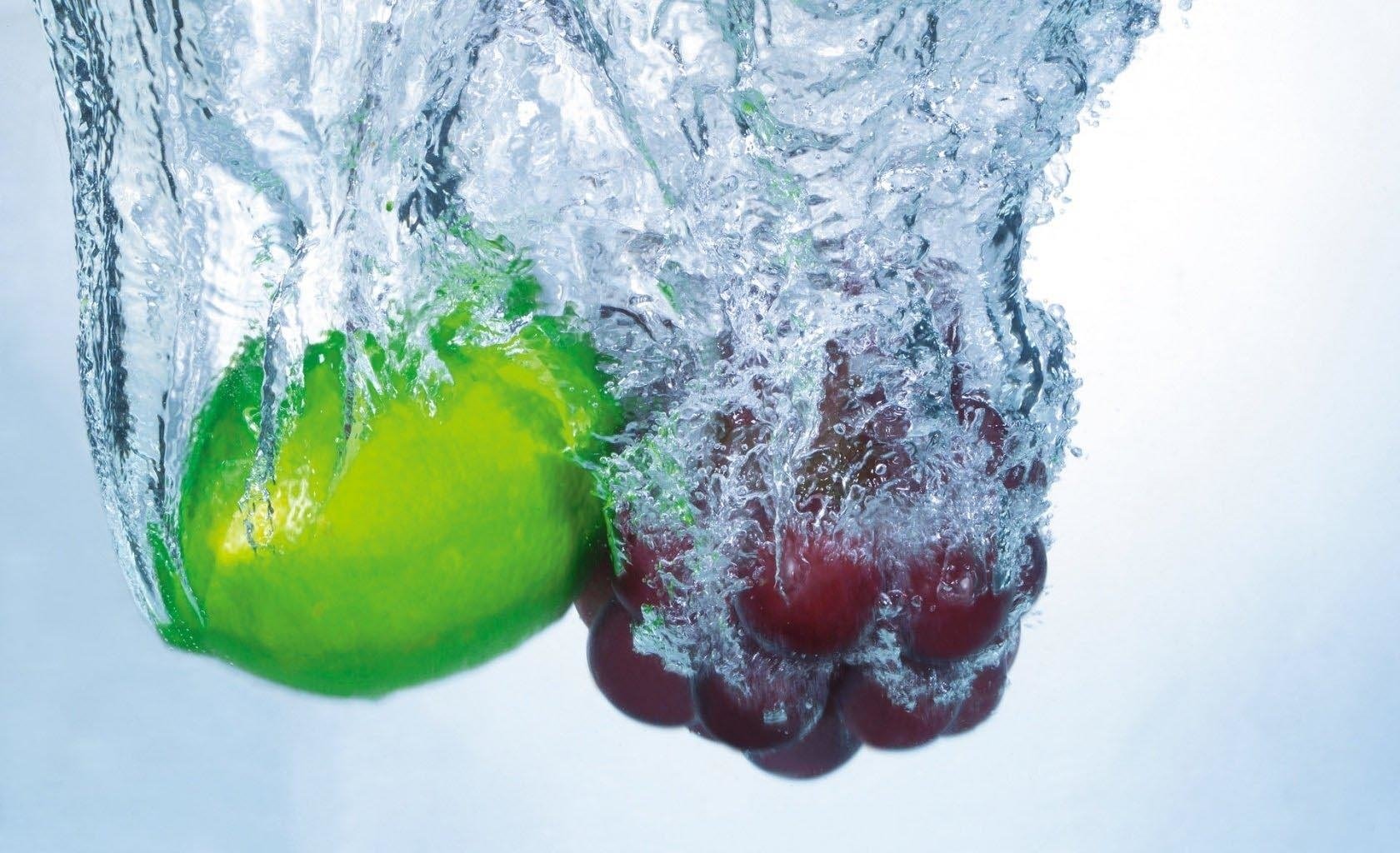 A green lime and a cluster of red grapes splash into clear water, creating dynamic ripples. This vibrant scene highlights the freshness of food and fruit.