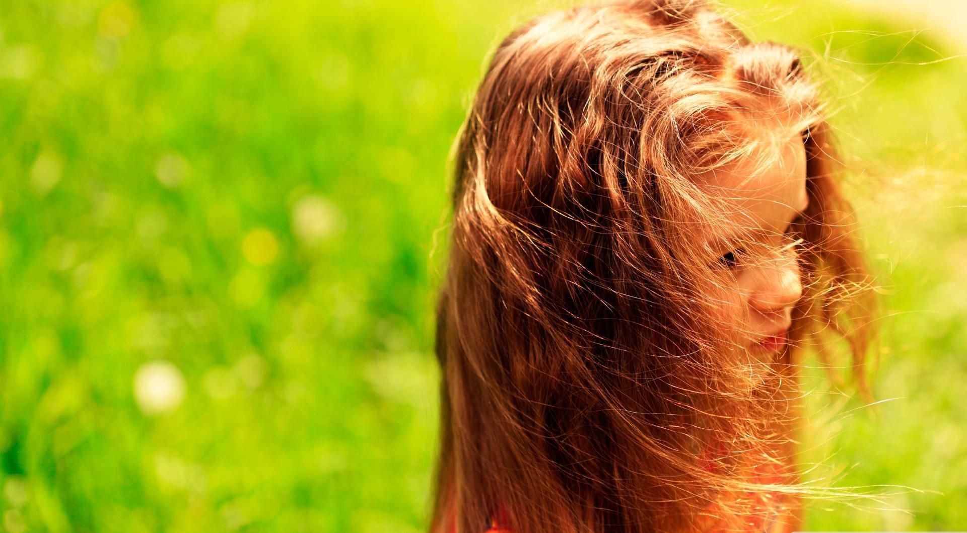 A child with long, flowing hair stands in a sunlit green field, looking thoughtfully to the side, embodying a moment of serene childhood wonder.