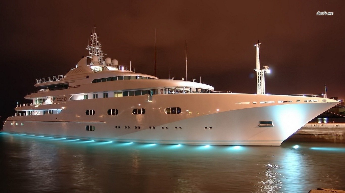 Night view of a large illuminated yacht, a marine vehicle moored at a waterfront, blue under-hull lights casting reflections on calm water.