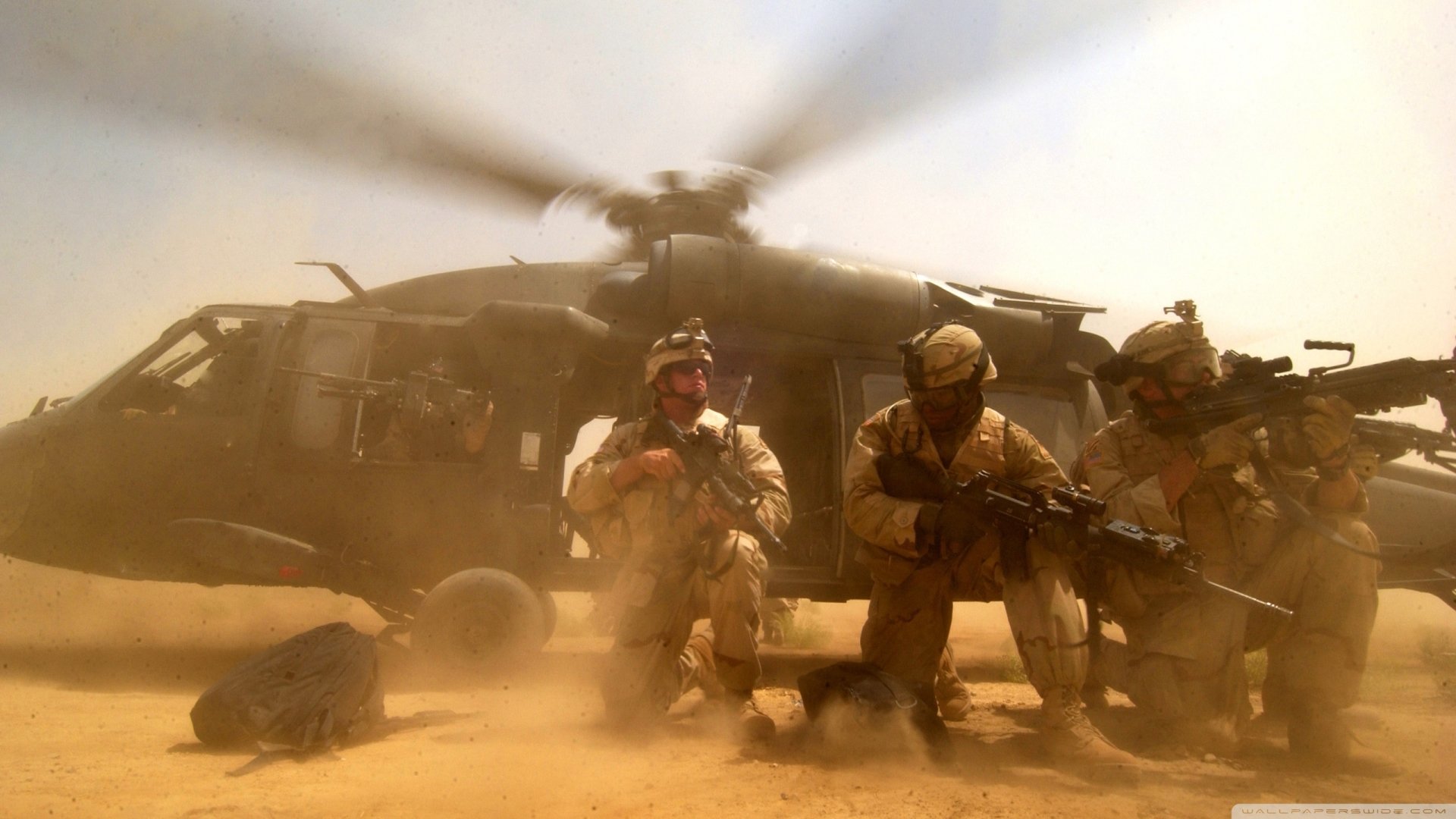Military soldiers in desert gear kneel and prepare weapons beside a dust-churned helicopter as sand blows around them.