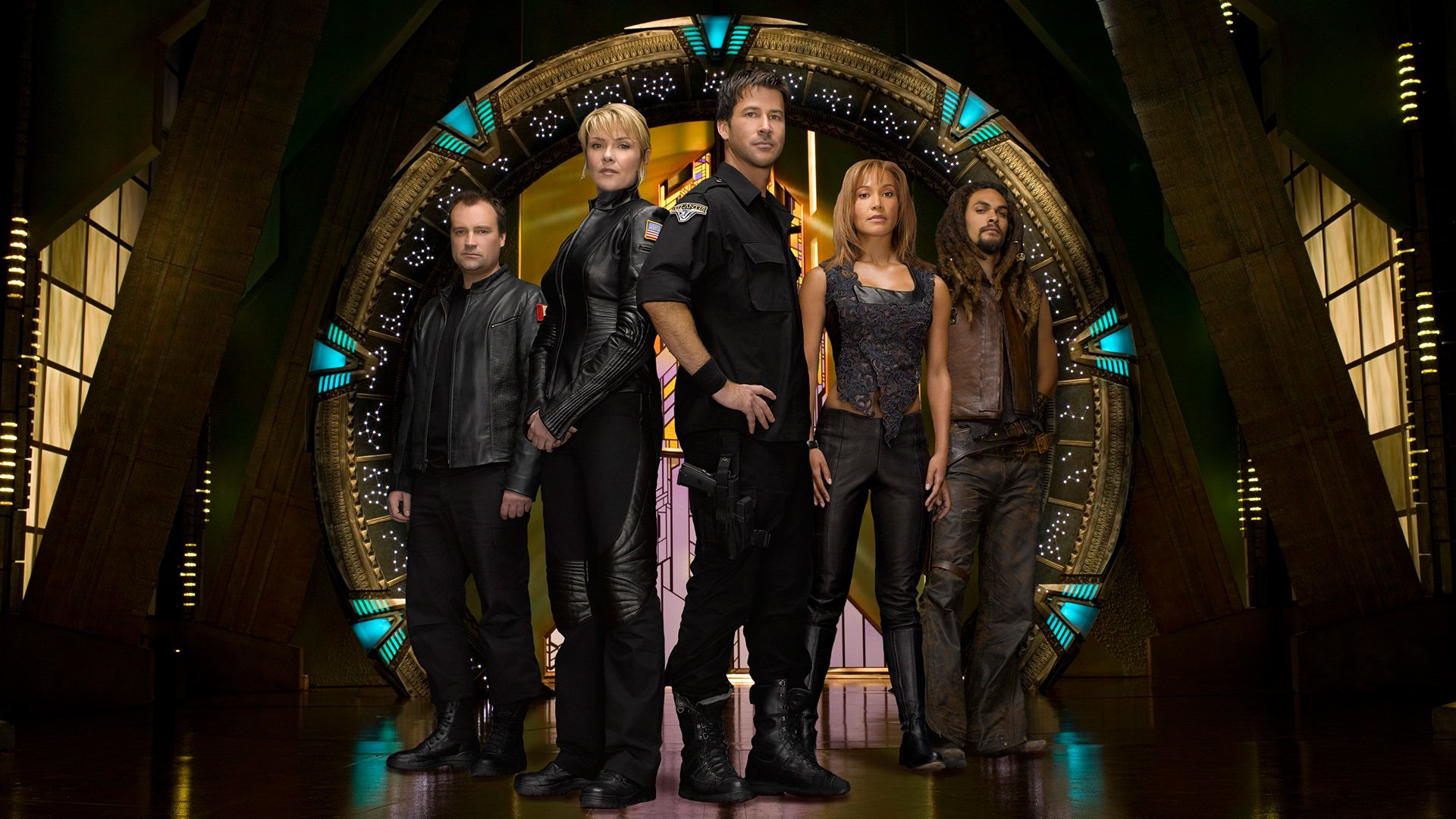 Five members of the TV show Stargate Atlantis cast stand before the activated Stargate, wearing military sci‑fi gear, framed by a glowing circular ring and metallic archway.