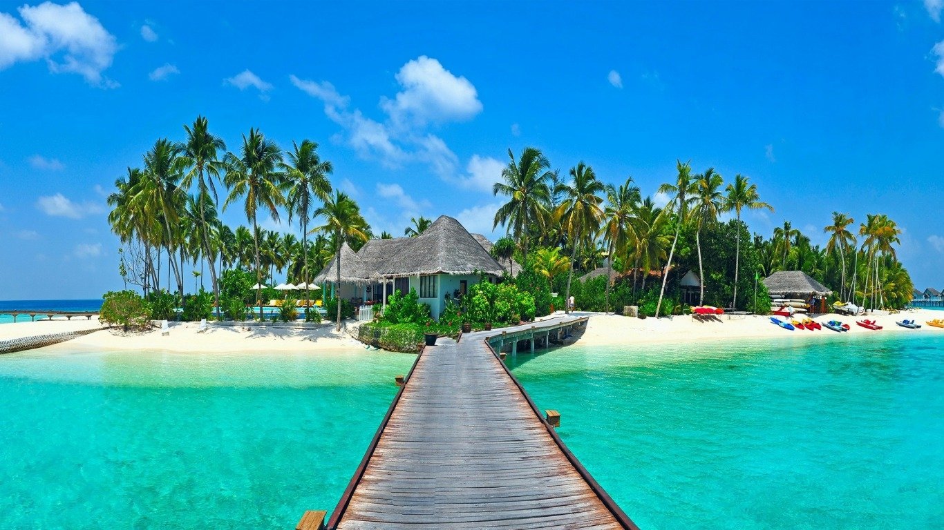 A tropical island with palm trees, clear turquoise water, and a wooden walkway leading to thatched-roof huts, captured in vibrant holiday photography.