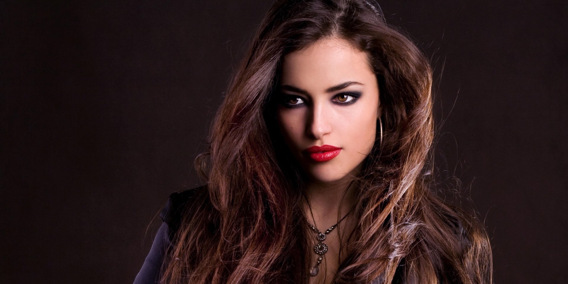 Close-up of a woman's face framed by long wavy hair, smoky eye makeup and bold red lips, gazing off to the side against a dark background.