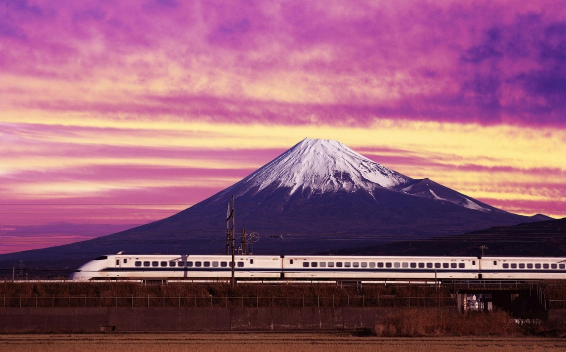 A sleek white train vehicle speeds past Mount Fuji, the snow-capped peak framed against a vibrant purple-orange sky.