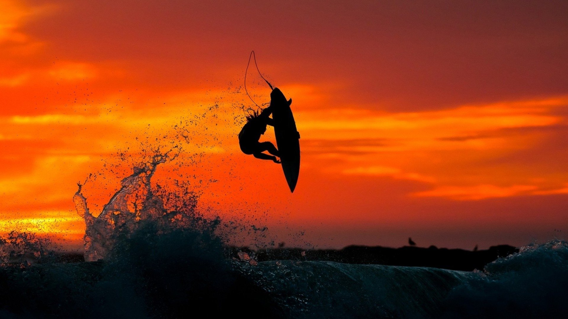 A surfer performs an aerial maneuver above crashing waves during a vibrant orange sunset, capturing the thrill of surfing and water sports.