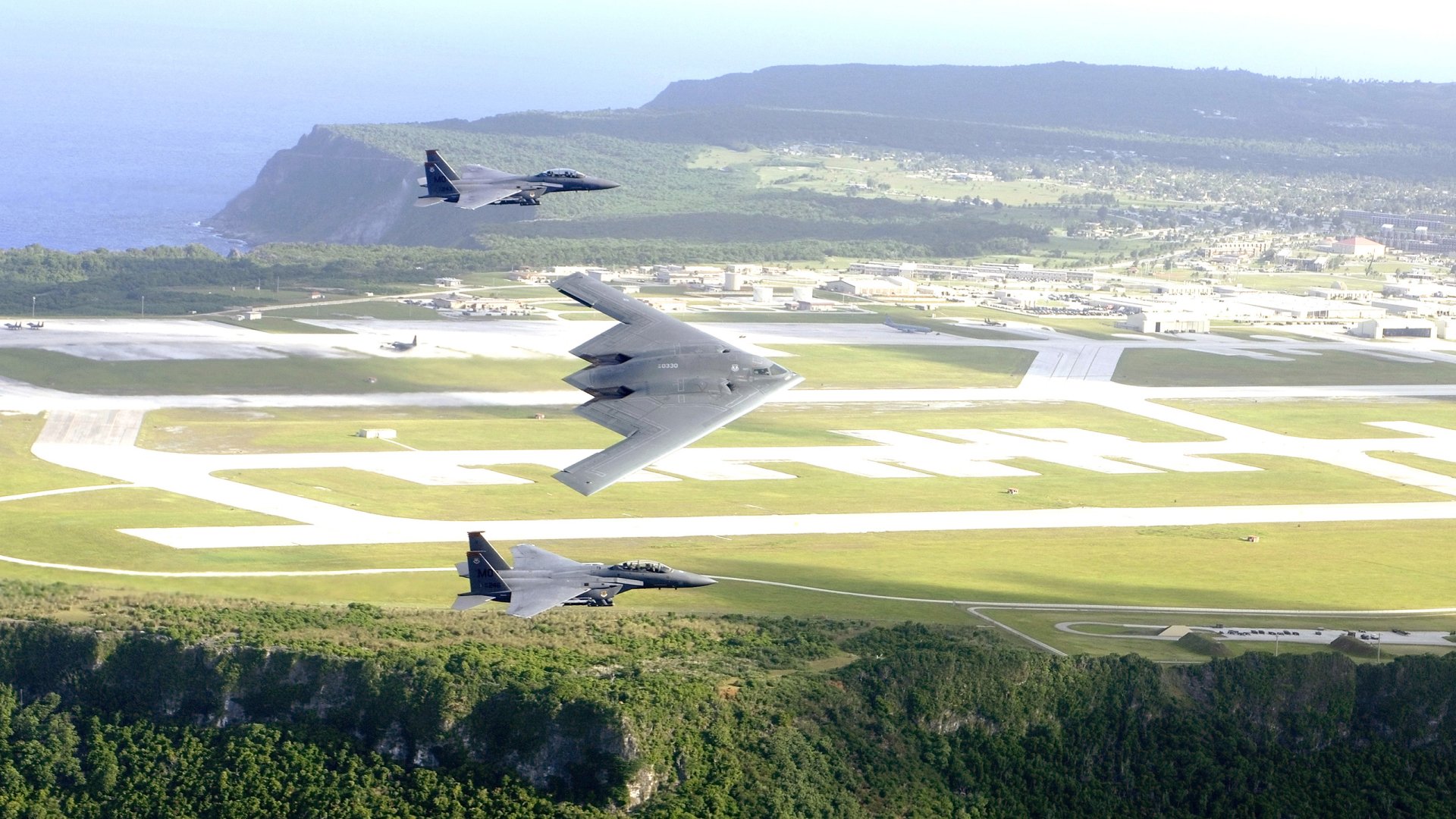 Three military aircraft fly in formation over a runway during an air show, with green hills and a distant coastline in the background.