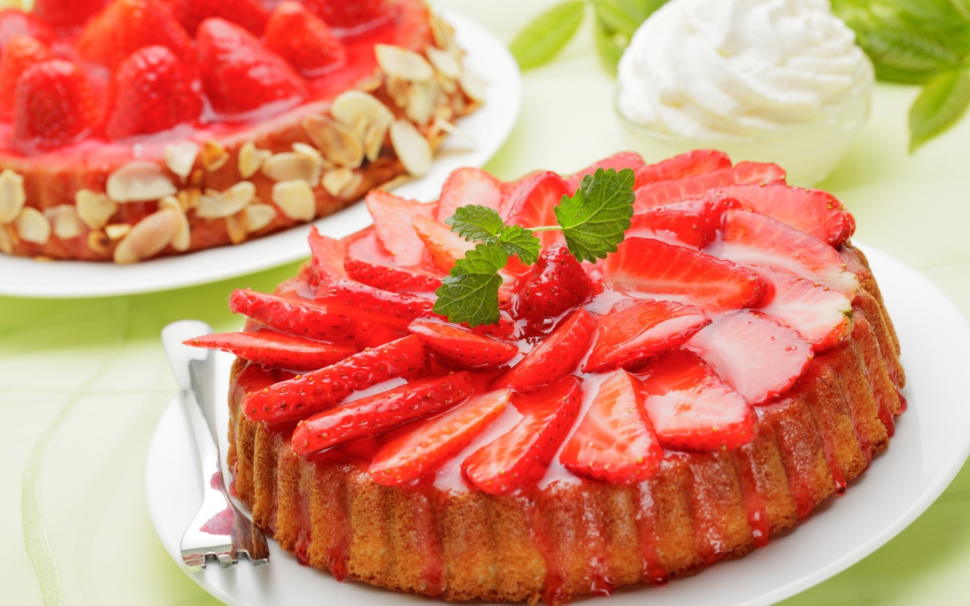A vibrant strawberry cake topped with fresh strawberries and a mint garnish, served alongside another cake with a glossy fruit glaze and almond slivers. A delightful display of desserts.