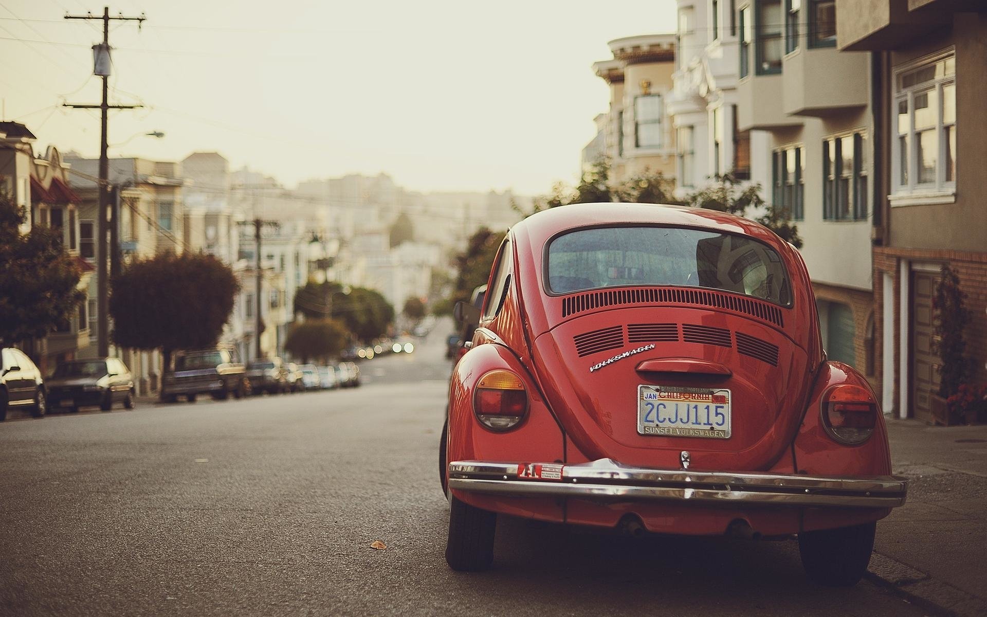 Red vintage Volkswagen Beetle parked on a sloped city street lined with houses in warm evening light.