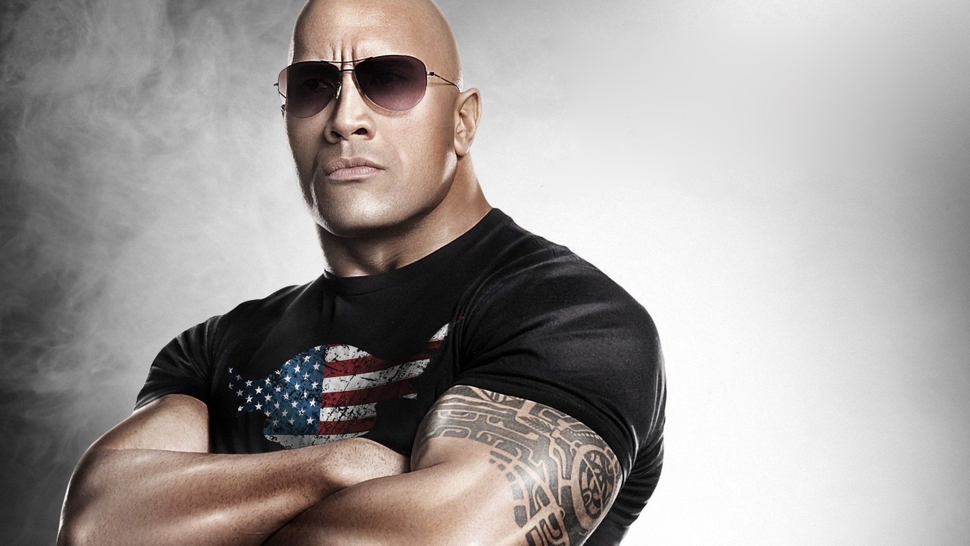 Dwayne Johnson poses confidently with crossed arms, wearing sunglasses and a black t-shirt featuring the American flag, against a dramatic light backdrop.