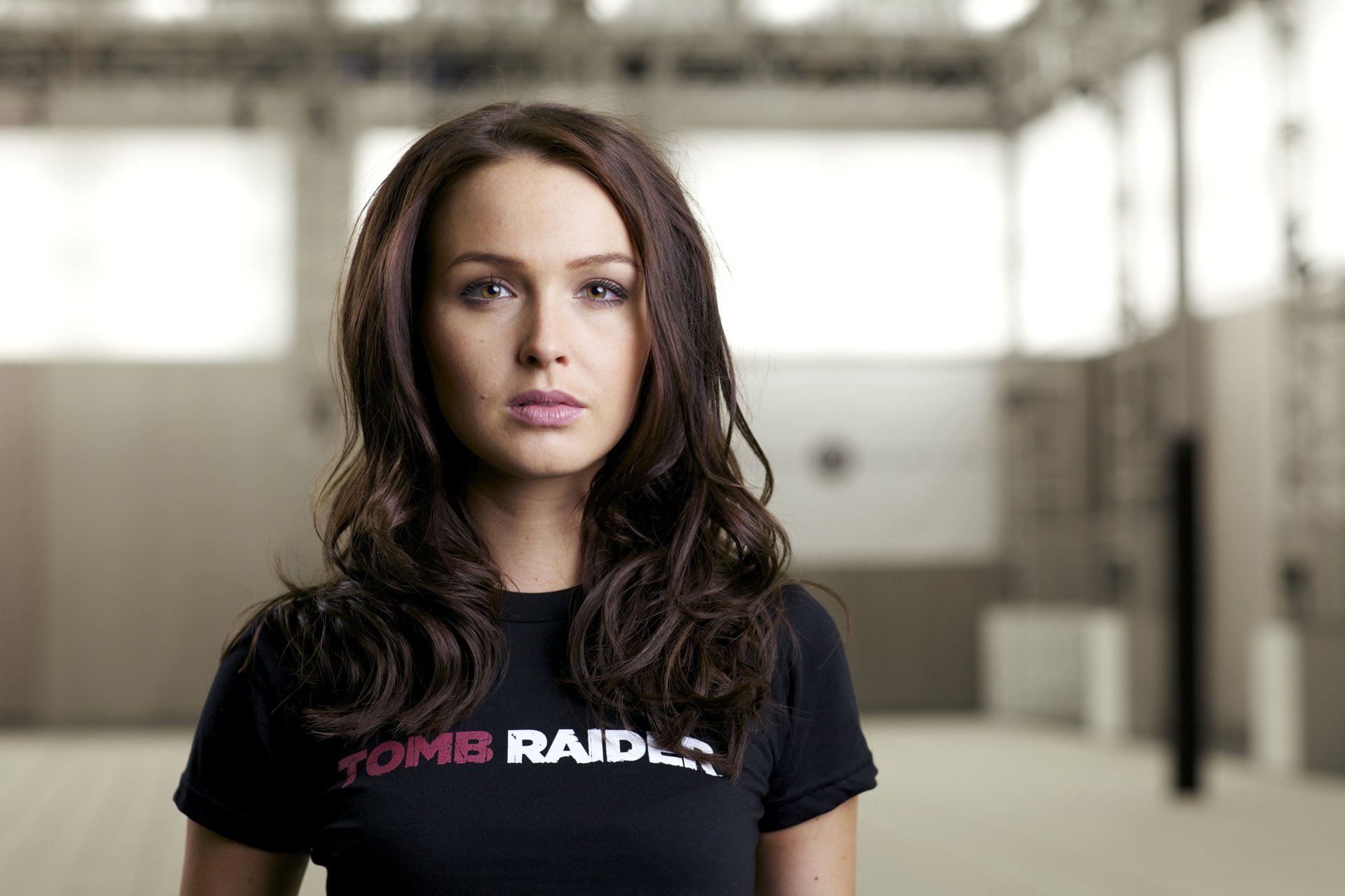 Portrait of woman Camilla Luddington with long dark hair, wearing a black Tomb Raider t-shirt, standing in a softly lit industrial space.