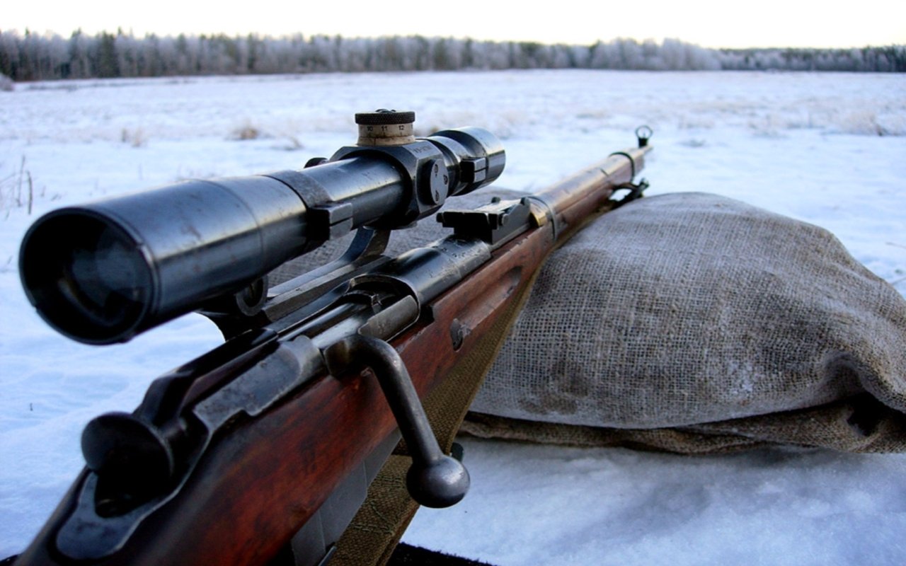 A close-up of a sniper rifle with a scope, resting on a burlap sack in a snowy landscape, showcasing the craftsmanship of this man-made firearm.