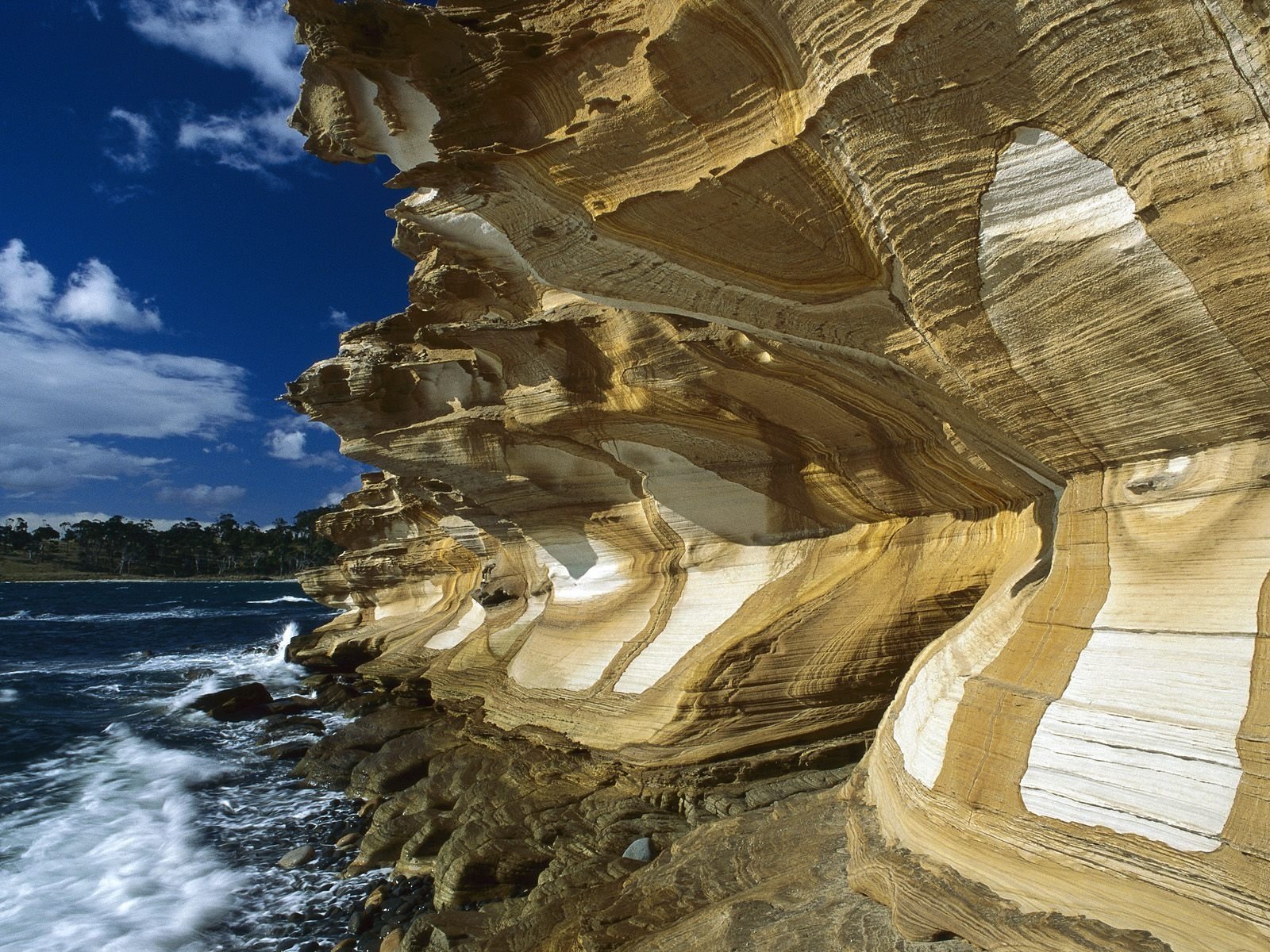 A rugged cliff showcasing layered rock formations meets the ocean, with waves crashing against the base under a bright blue sky dotted with clouds.