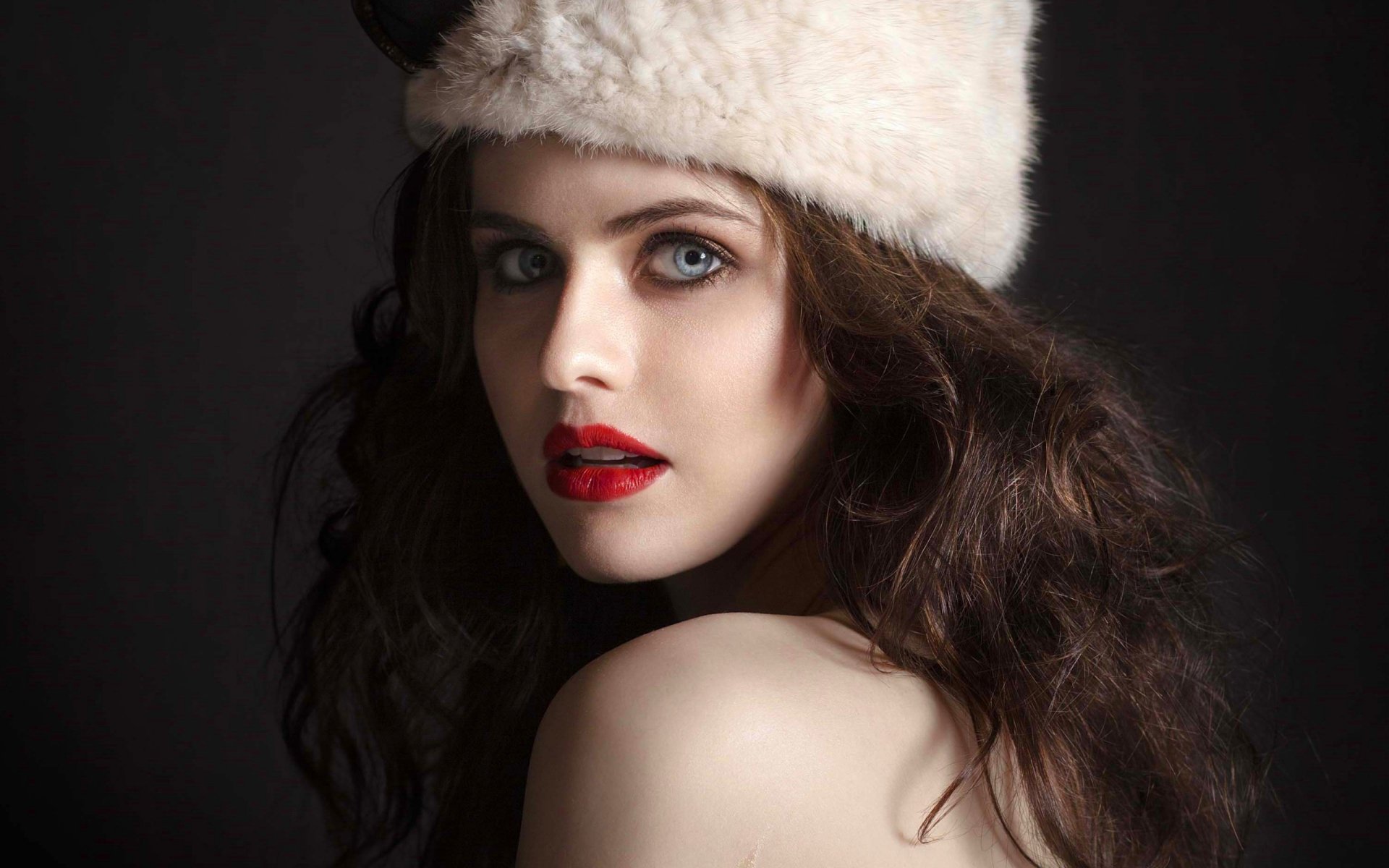 Portrait of celebrity Alexandra Daddario wearing a white fur hat, with long dark hair, striking blue eyes, and red lipstick against a dark background.
