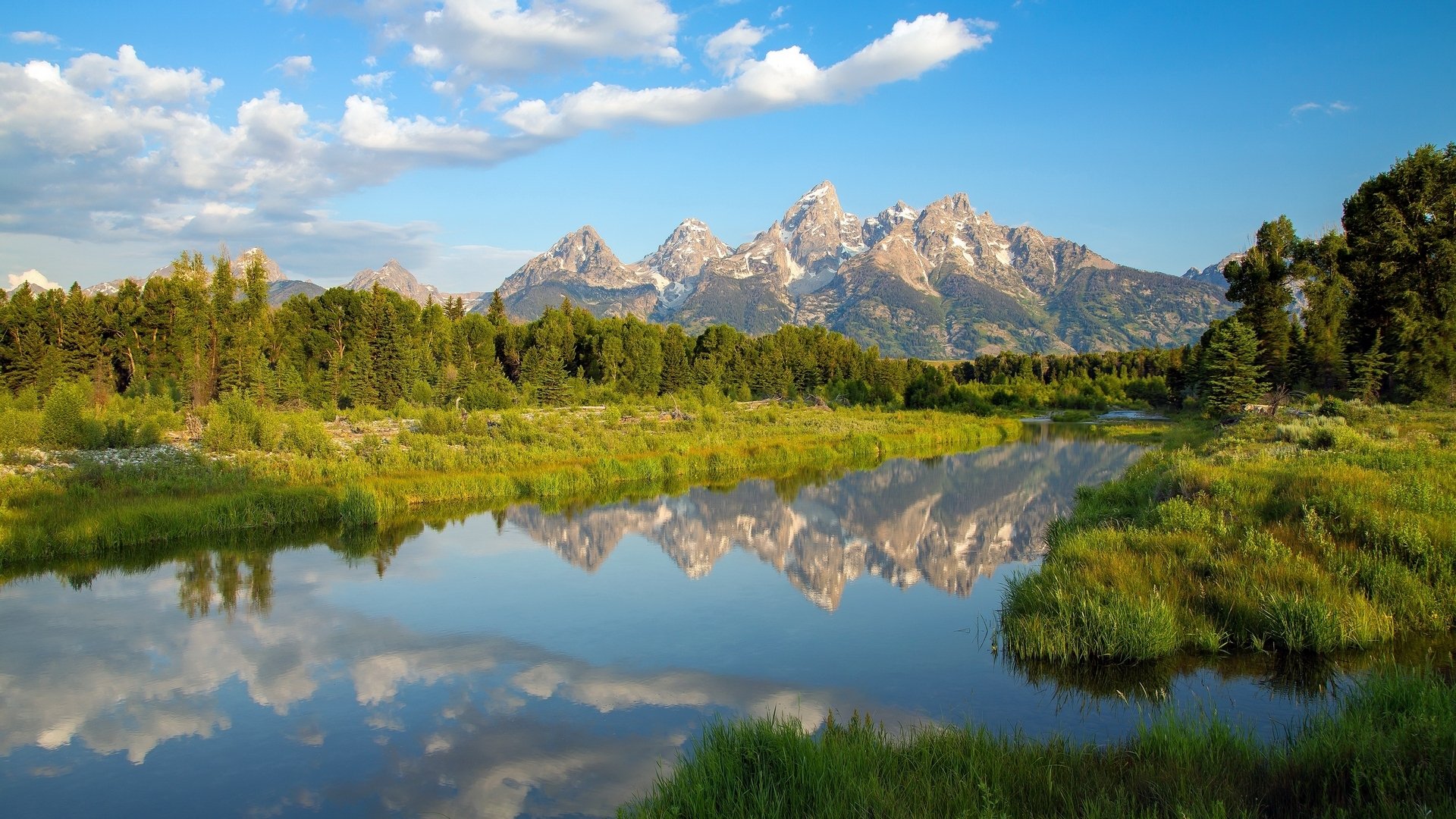 A tranquil nature scene with mountains and trees reflected perfectly in a calm river under a bright blue sky with scattered clouds.
