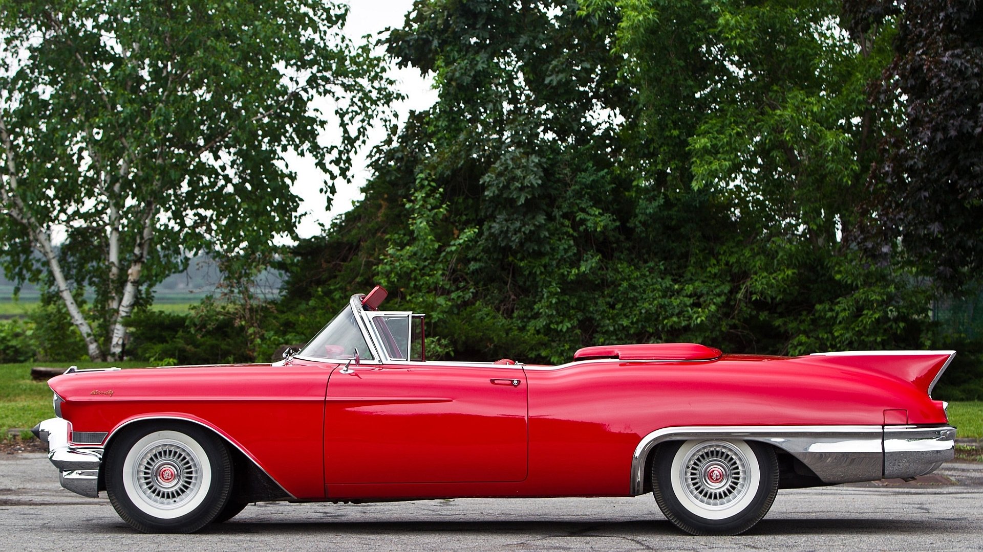 A bright red Cadillac Eldorado Biarritz convertible parked on a road with trees and greenery in the background.