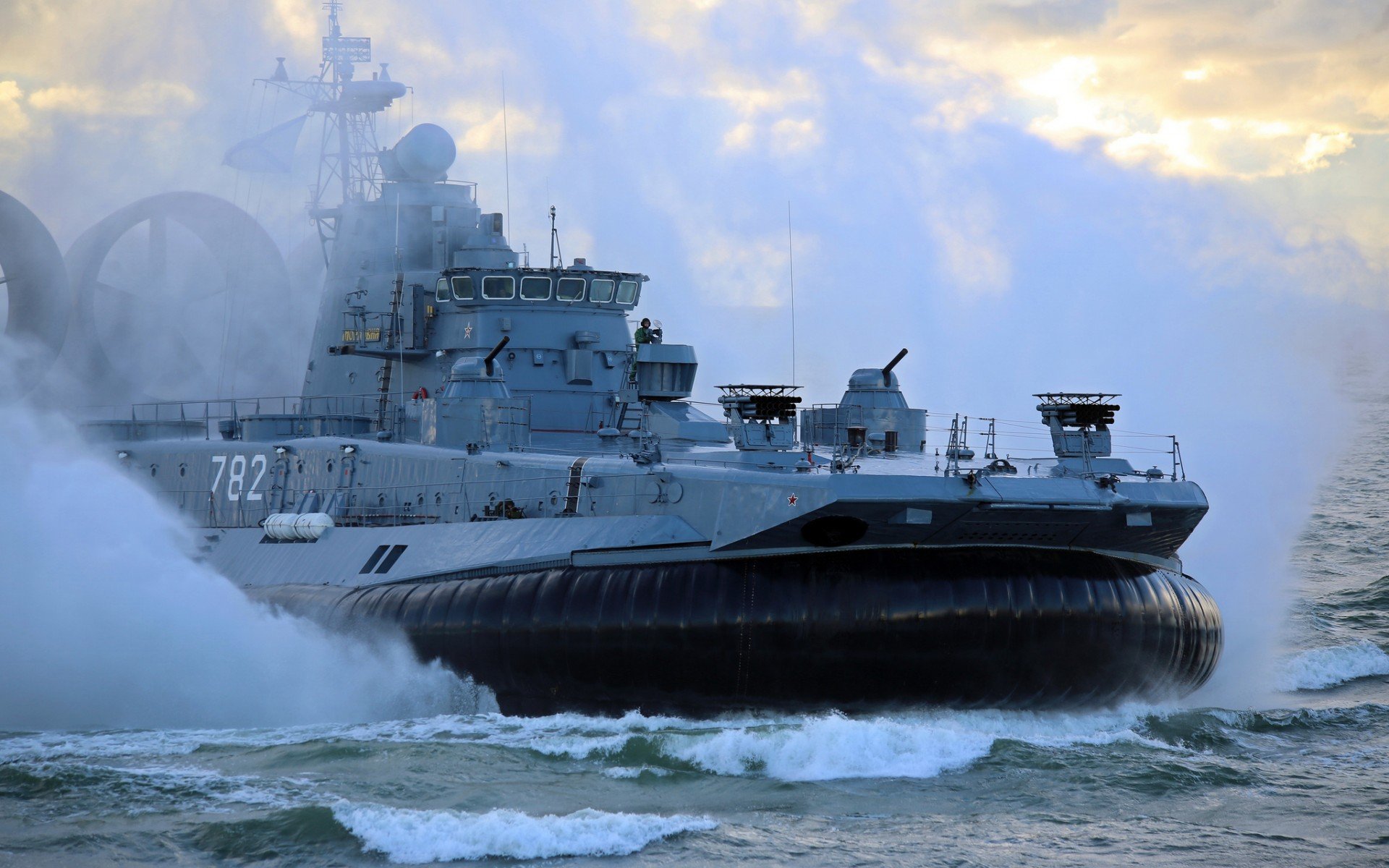 A military hovercraft glides over turbulent waters, creating a splash as it navigates through the waves under a cloudy sky.